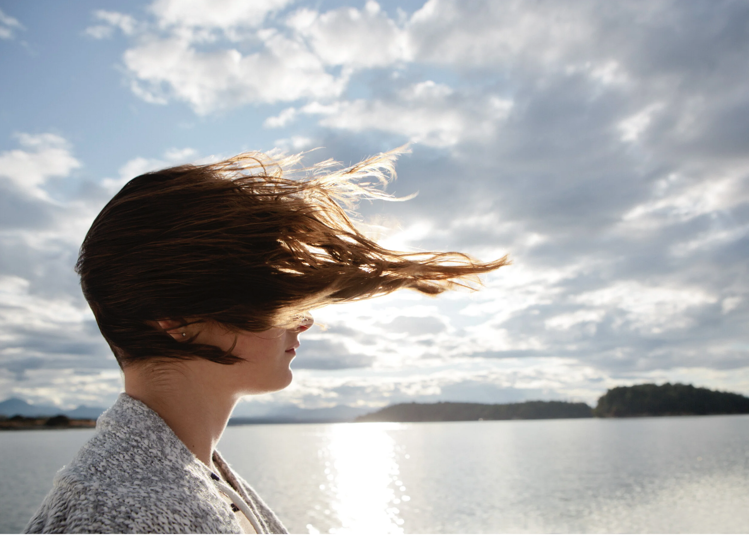A woman with short brown hair looking to the side outdoors near a body of water, with cloudy sky and distant trees in the background.