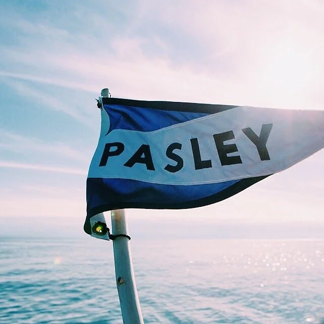 A blue and white flag with the word "PASLEY" flying over water during sunset or sunrise.