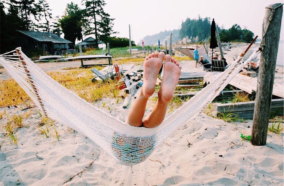 Person lying in a hammock at the beach with their legs and feet hanging out over the edge, facing away from the camera. The beach scene includes some chairs, umbrellas, and a rustic shoreline with trees and houses in the background.