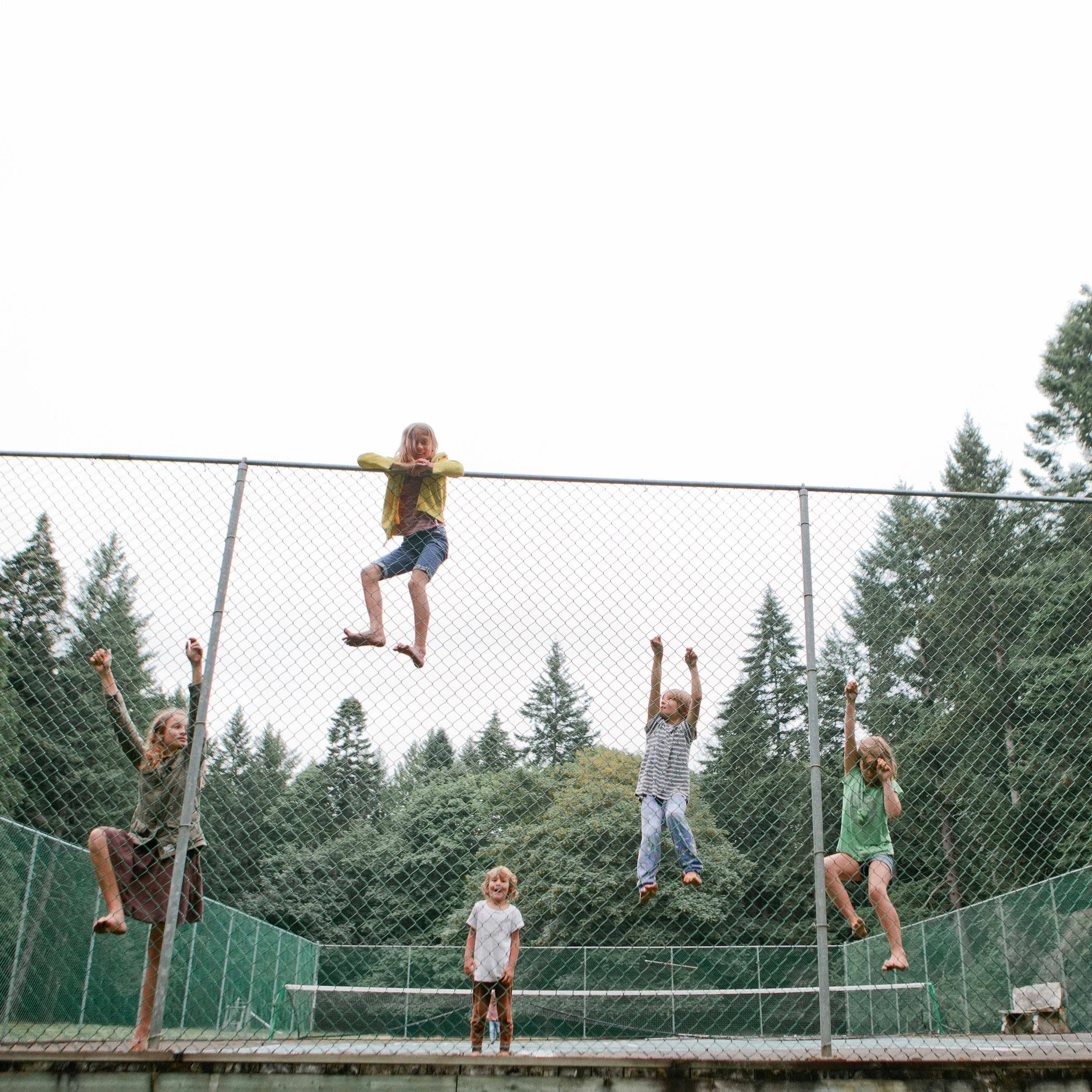 Young girl on a trampoline with safety net, surrounded by trees and cloudy sky, enjoying a sunny day.
