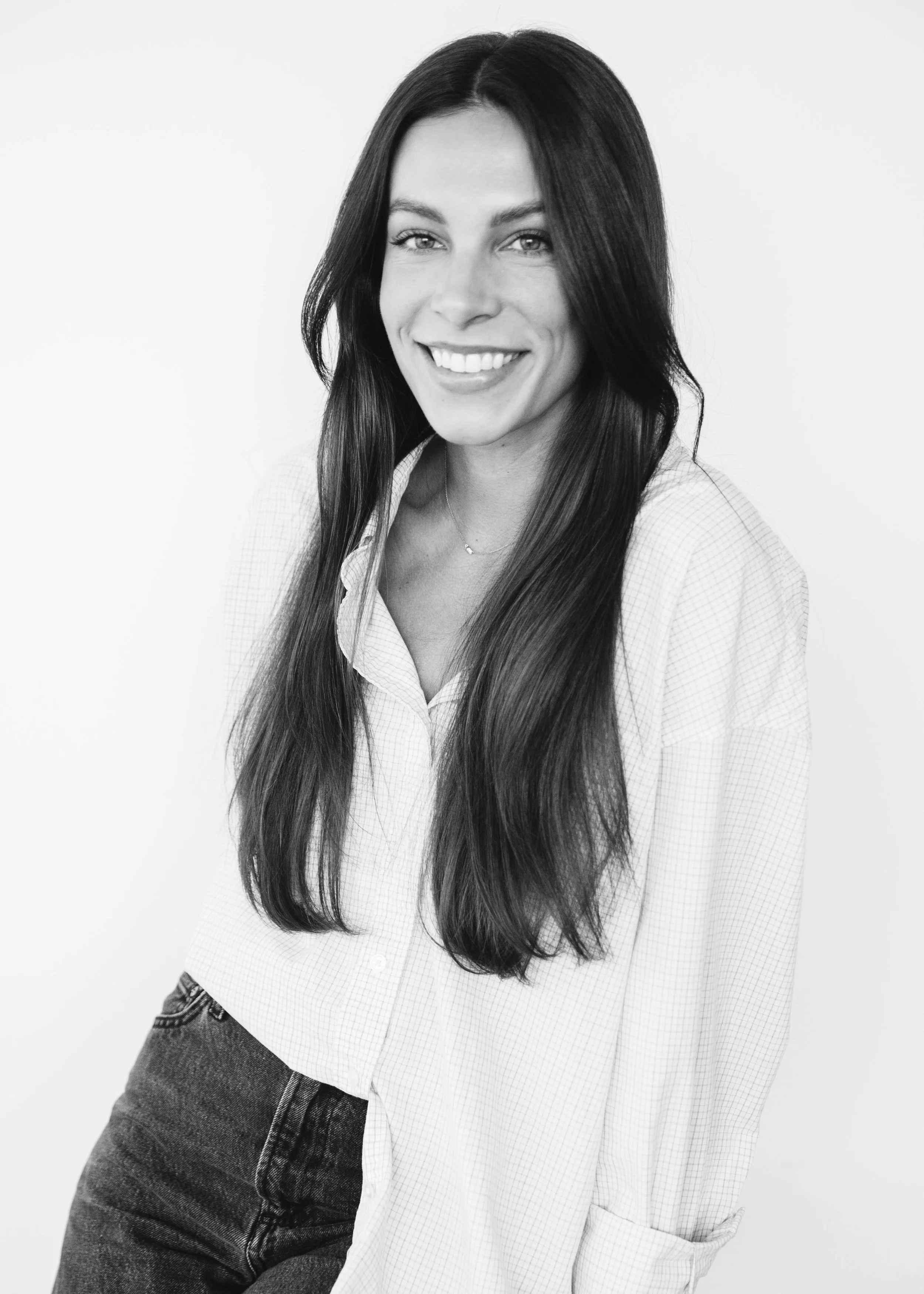 Black and white portrait of a smiling woman with long hair, wearing a checkered shirt and dark jeans, standing against a plain background.