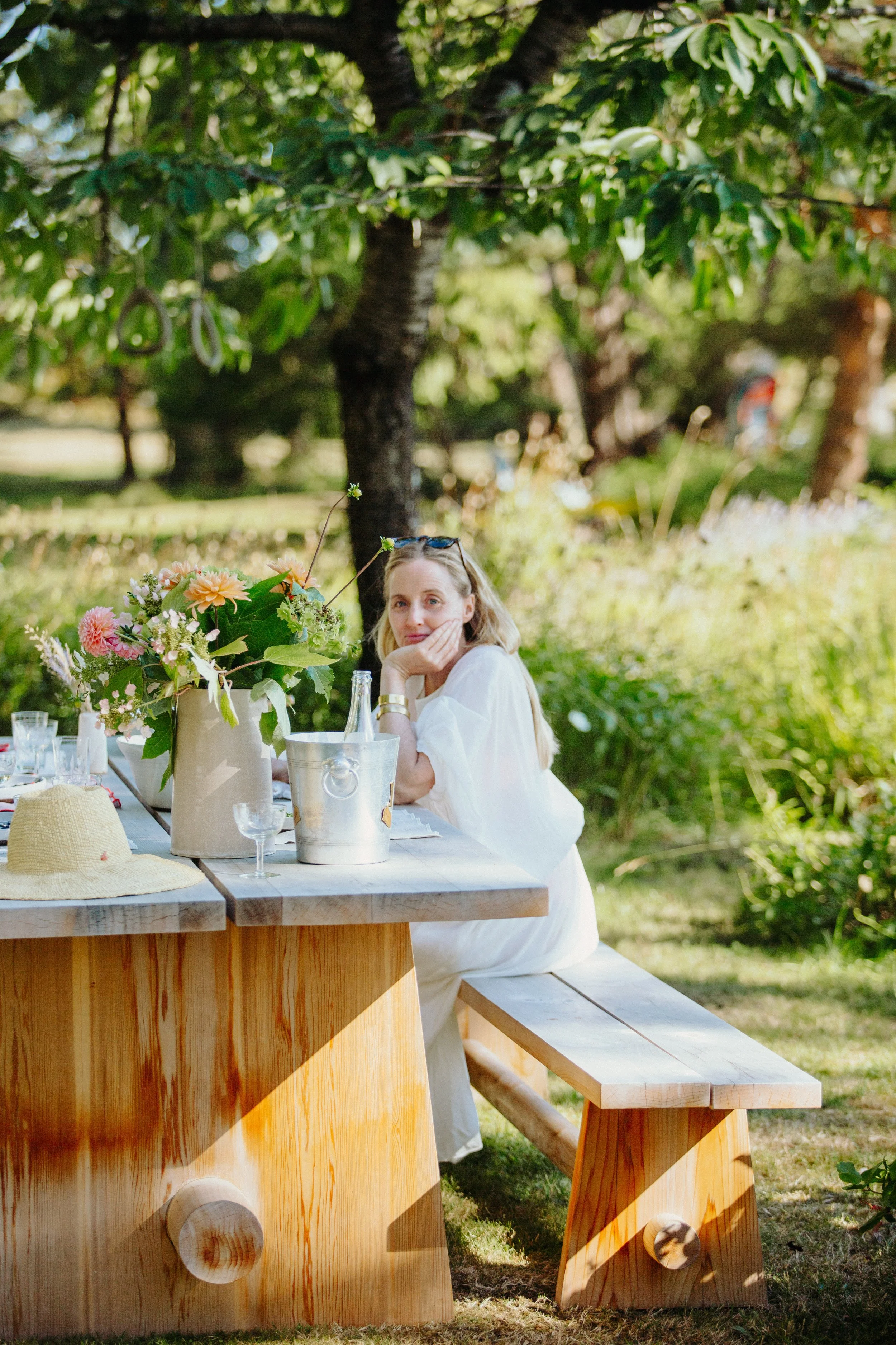 A woman in a white dress sitting at an outdoor wooden table decorated with a large floral arrangement, a hat, and glassware, in a lush green garden with trees and sunlight.