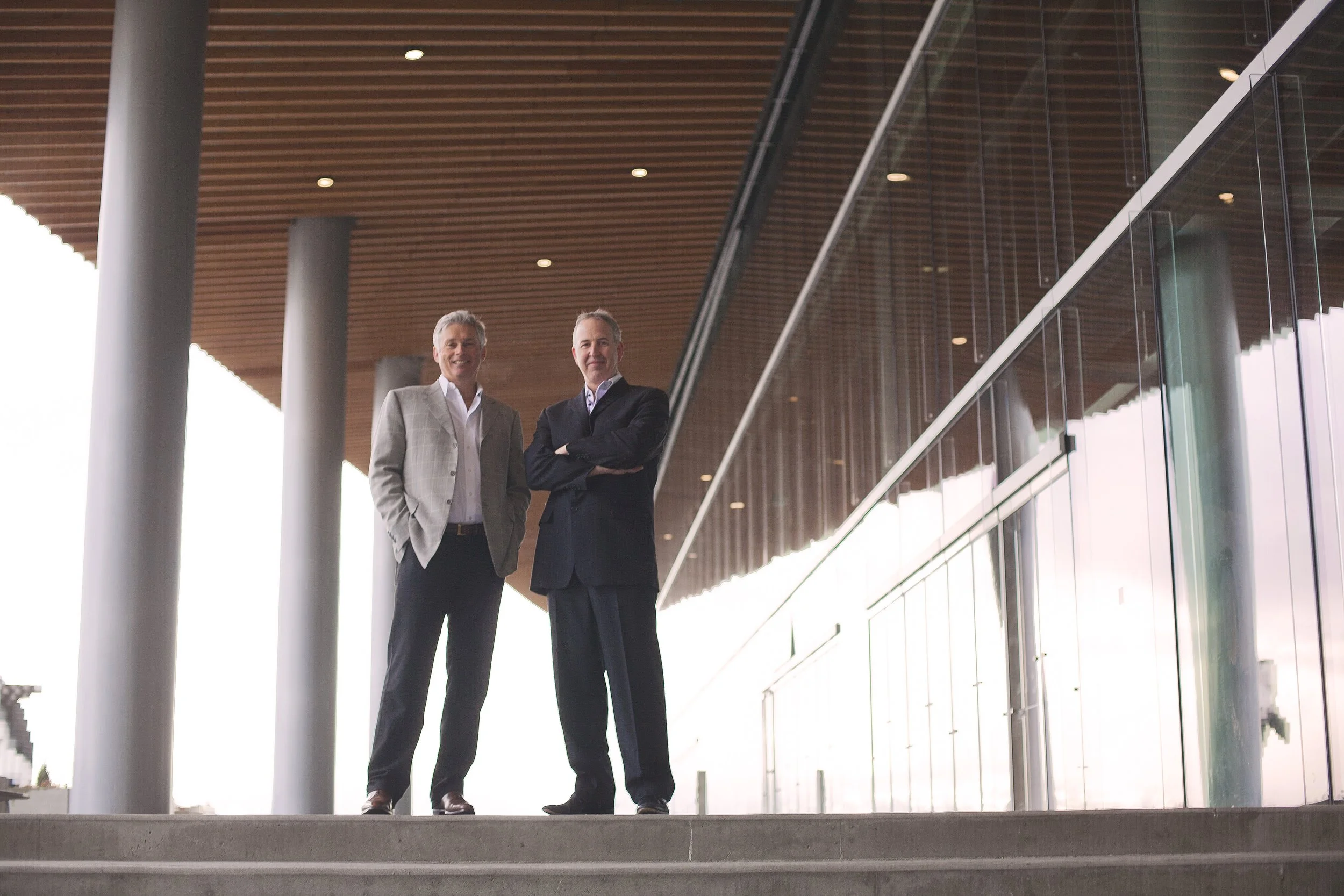Two middle-aged men in suits standing together in a modern building with a wooden ceiling and glass walls, smiling and posing for the photo.