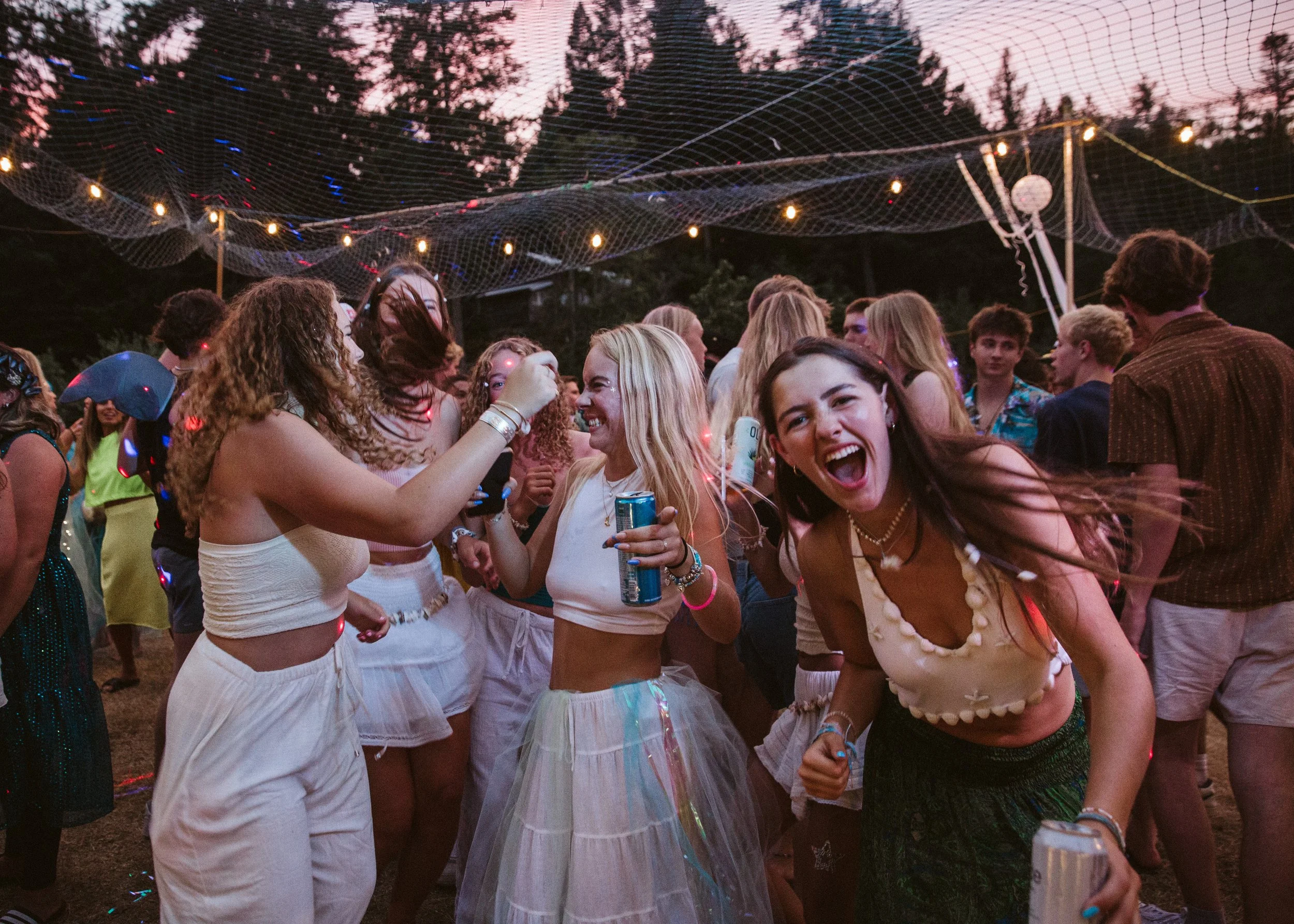 Group of young people dancing and enjoying at an outdoor party during sunset, with string lights overhead and trees in the background.