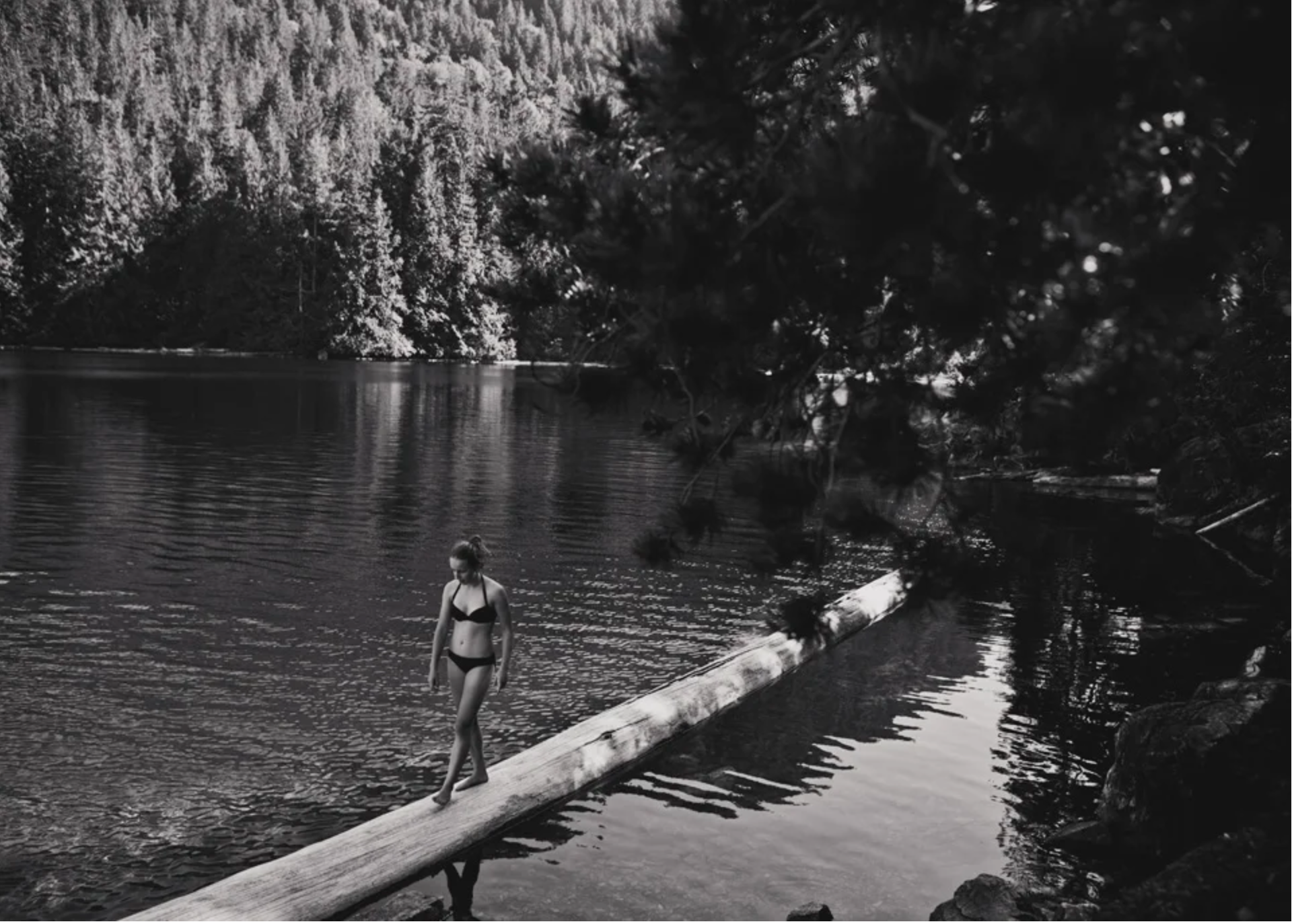 A person in a swimsuit walking on a fallen tree trunk across a calm river, with dense trees and a forested mountain in the background.