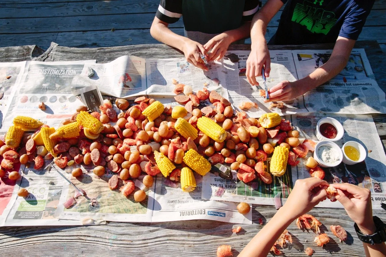 People preparing seafood boil with corn, potatoes, and sausage on a newspaper-covered wooden table.