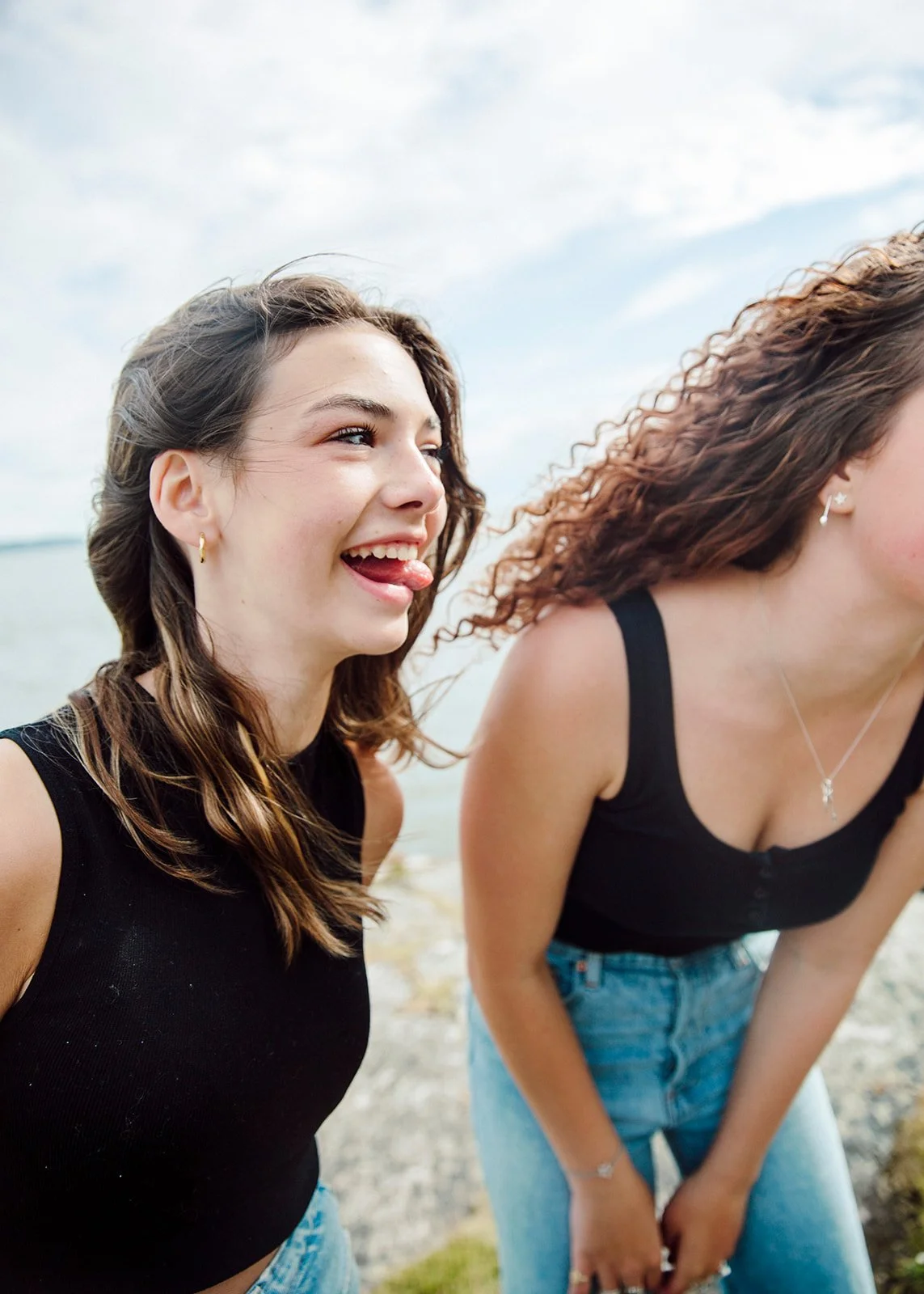 Two young women at the beach, one sticking out her tongue and smiling, with the ocean and cloudy sky in the background.