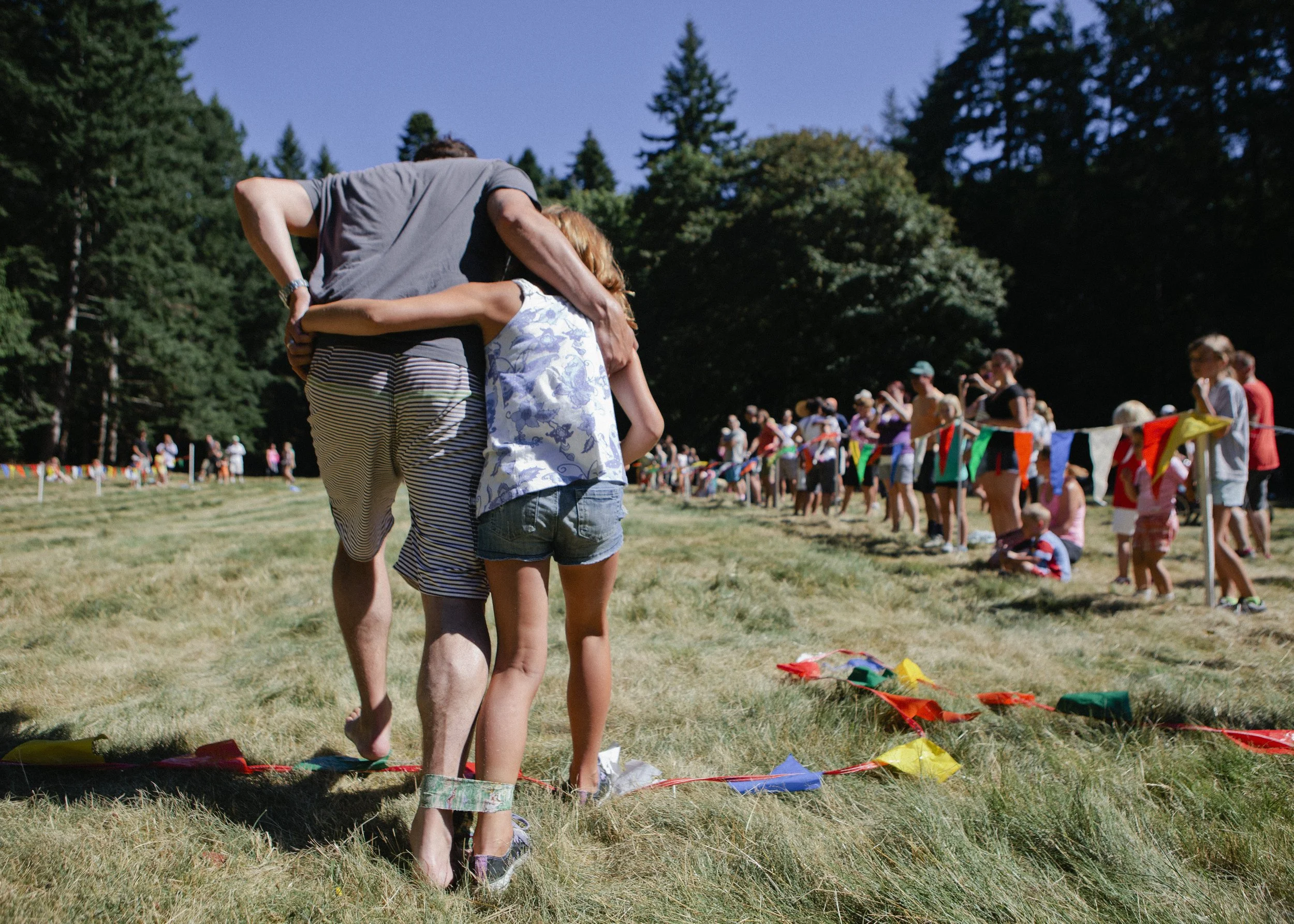 People lining up outdoors for an event, with trees and flags in the background on a sunny day.