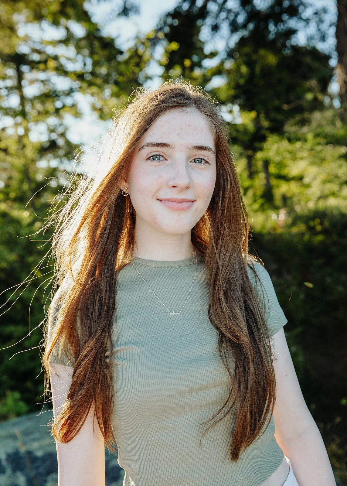 A young woman with long red hair, blue eyes, and freckles, smiling outdoors with green trees in the background. She is wearing a light green ribbed t-shirt, a silver necklace, and small hoop earrings, with sunlight illuminating her face.