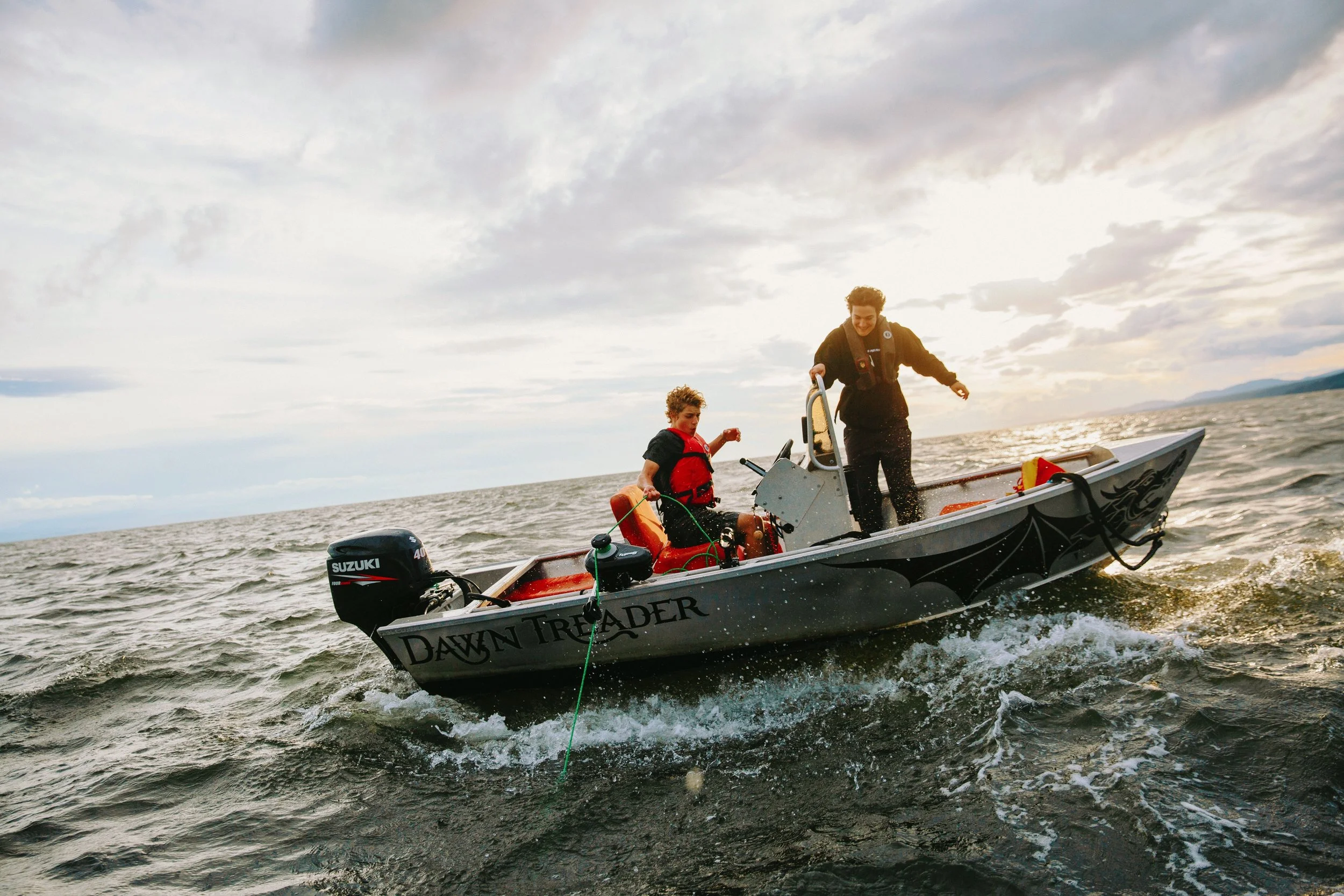 Two people on a small motorboat named Dawn Treader on the water, one standing and one sitting, with a cloudy sky and distant land in the background.