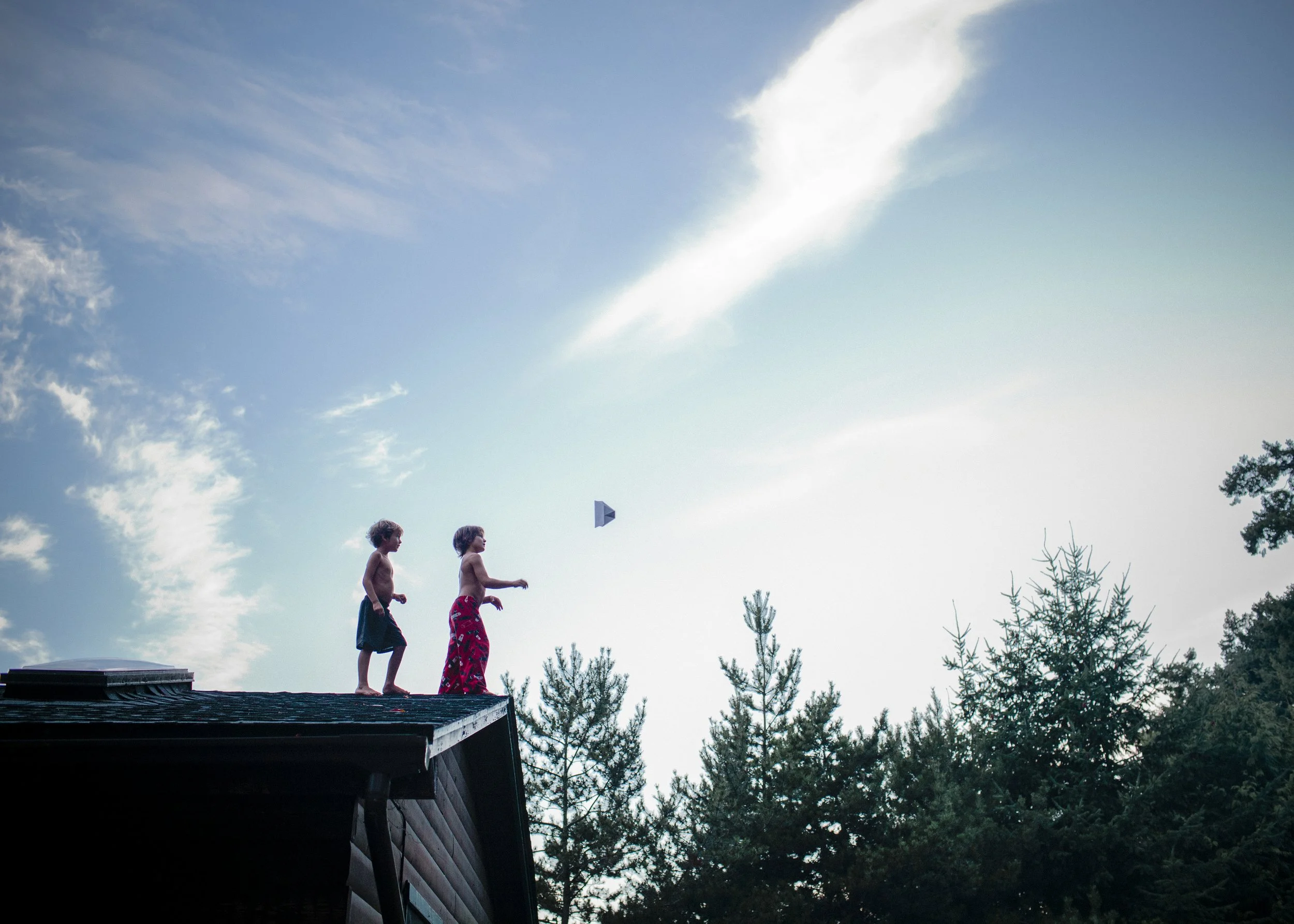 Two children, shirtless, walking on a roof during daytime, with trees and a partly cloudy sky in the background.
