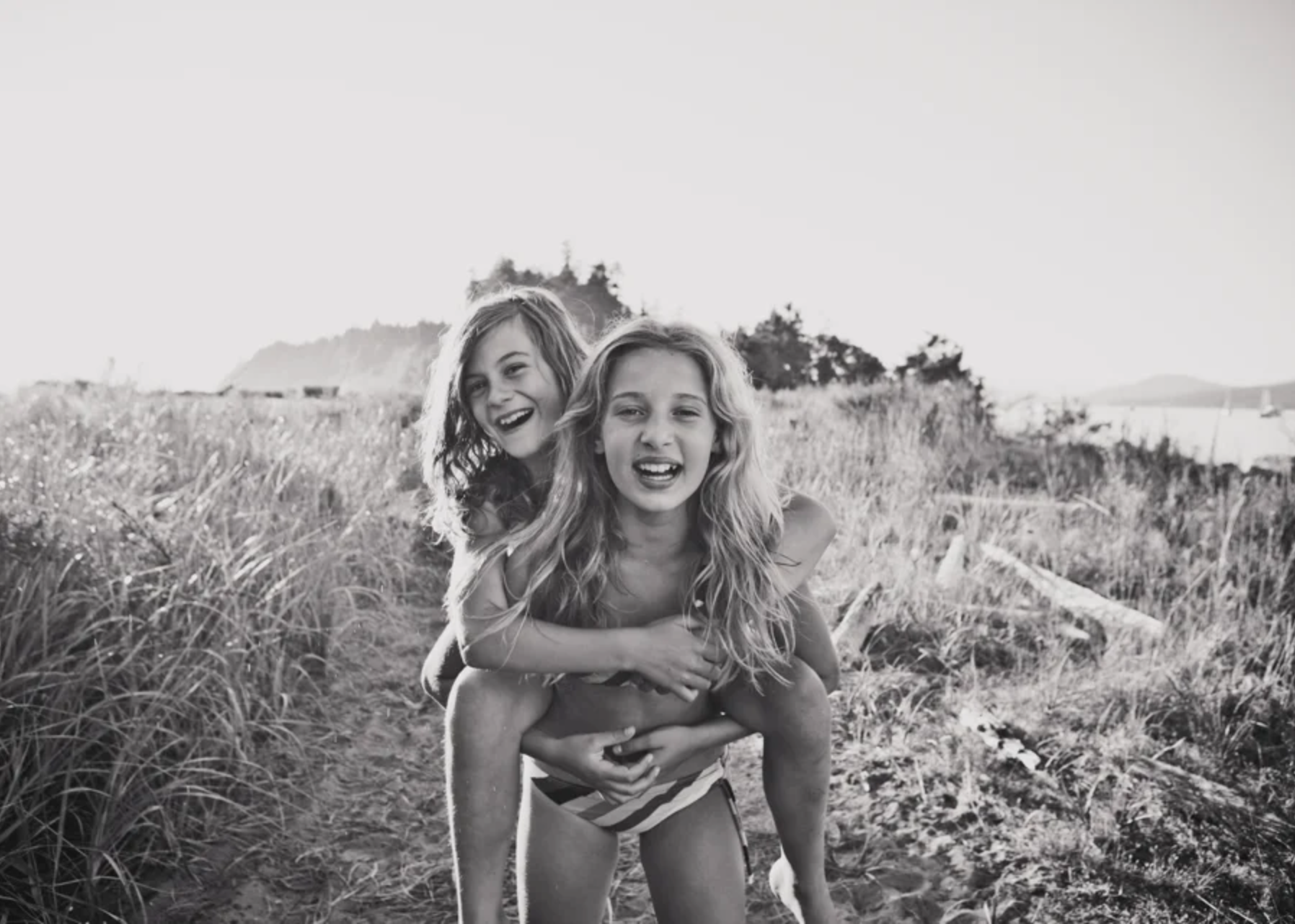 Two young girls playing at the beach, one giving the other a piggyback ride, with a scenic shoreline and trees in the background, in black and white.