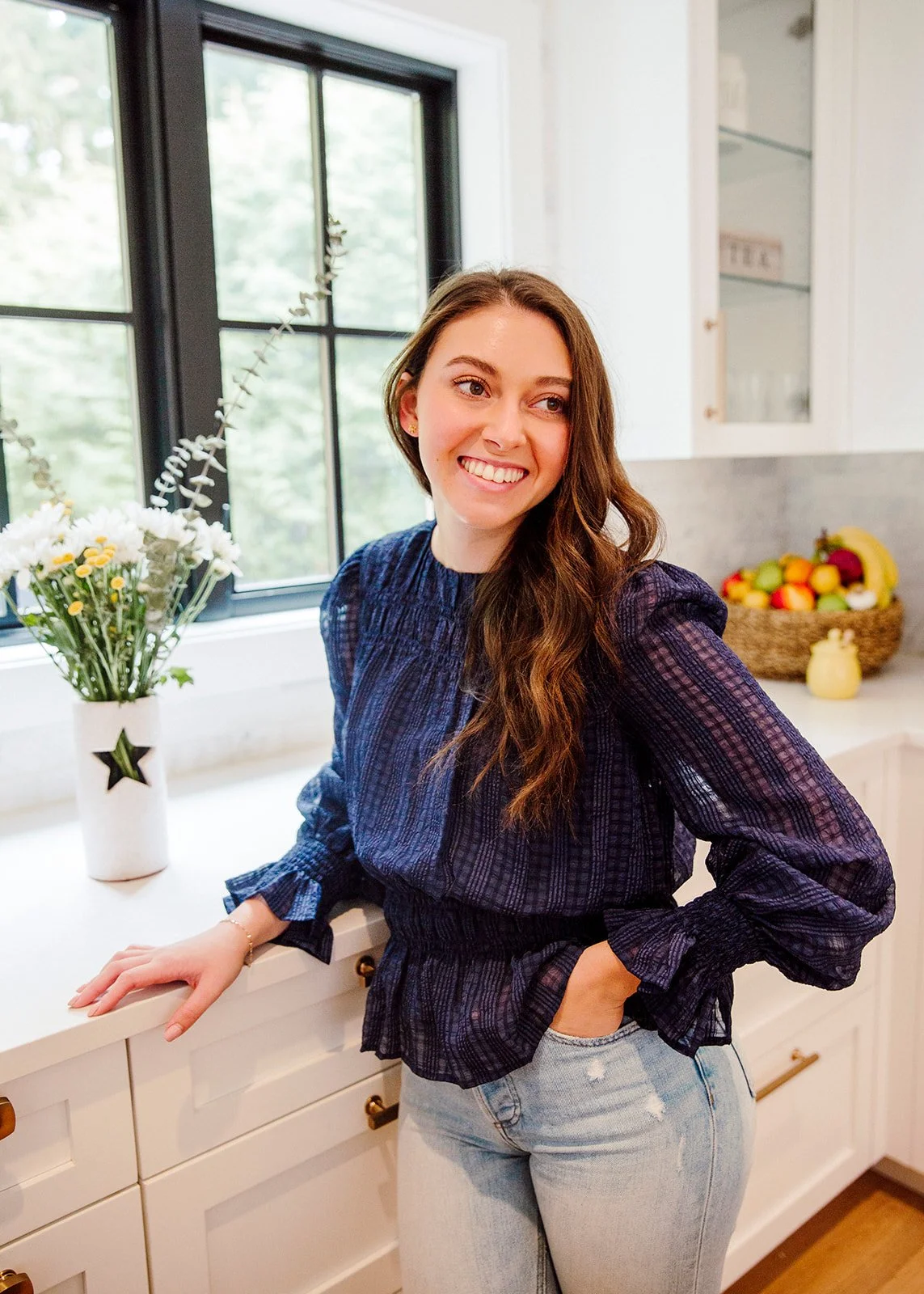 A young woman smiling and leaning on a white kitchen counter with a vase of flowers, a window behind her showing trees, and a bowl of fruit on the counter.