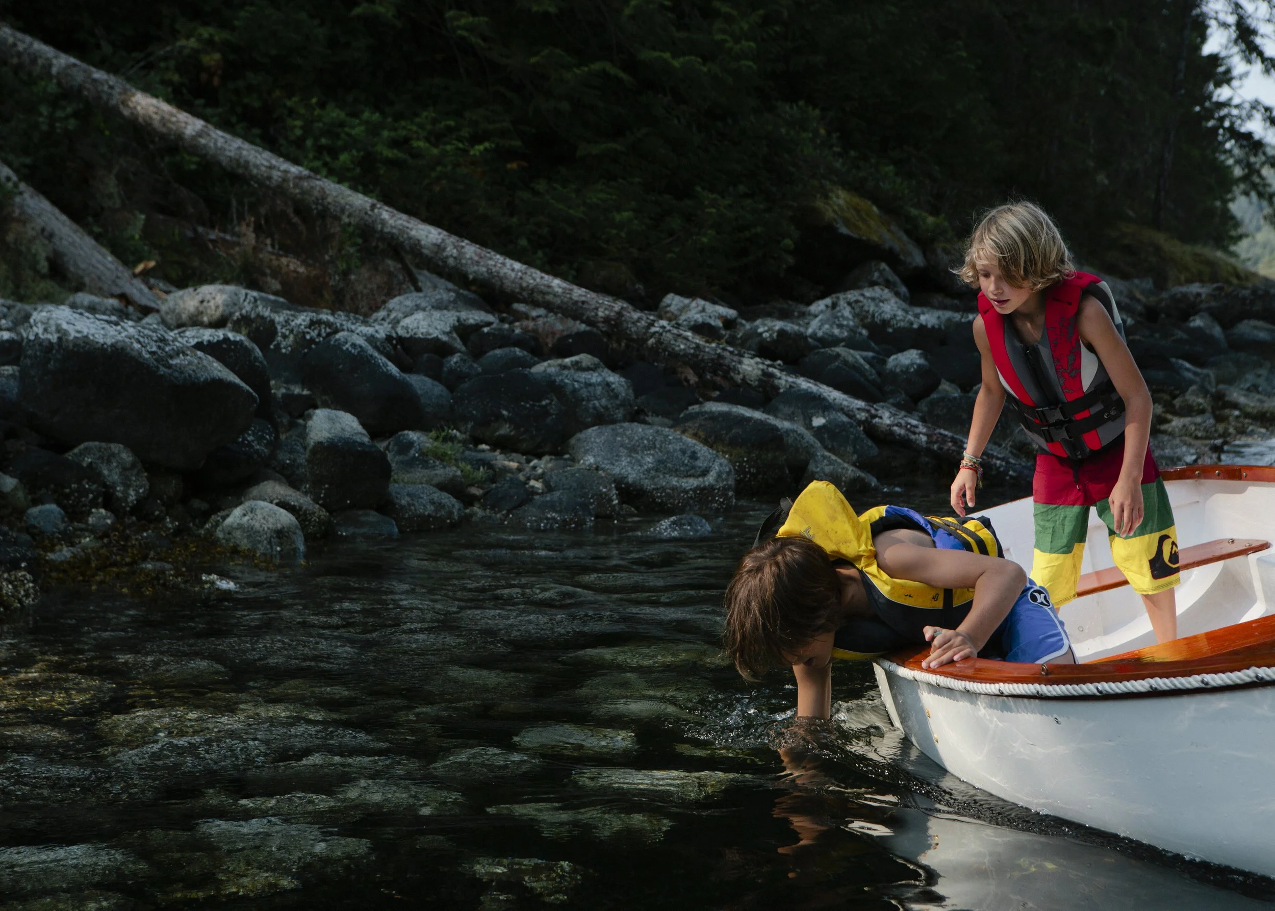 Two children on a small boat by a rocky shoreline, surrounded by trees and nature, wearing life jackets and colorful clothing.