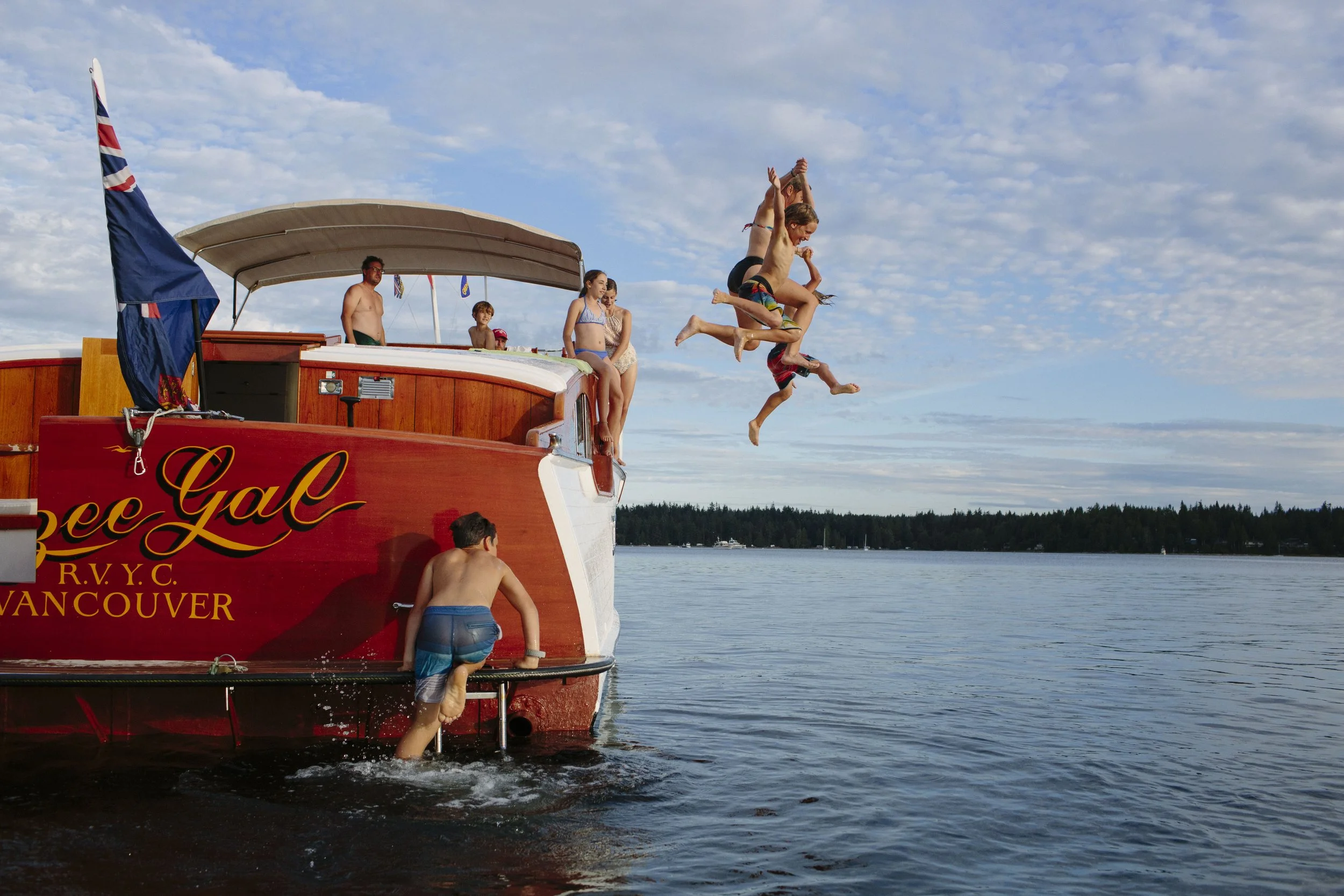 People jumping off a red boat into the water on a sunny day, with a blue sky and distant shoreline.