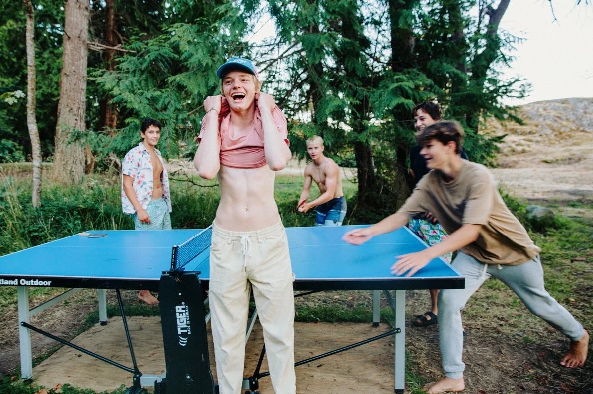 Group of young men playing table tennis outdoors in a forested area, one is laughing with a towel around his neck, others are reaching for the ball and watching.