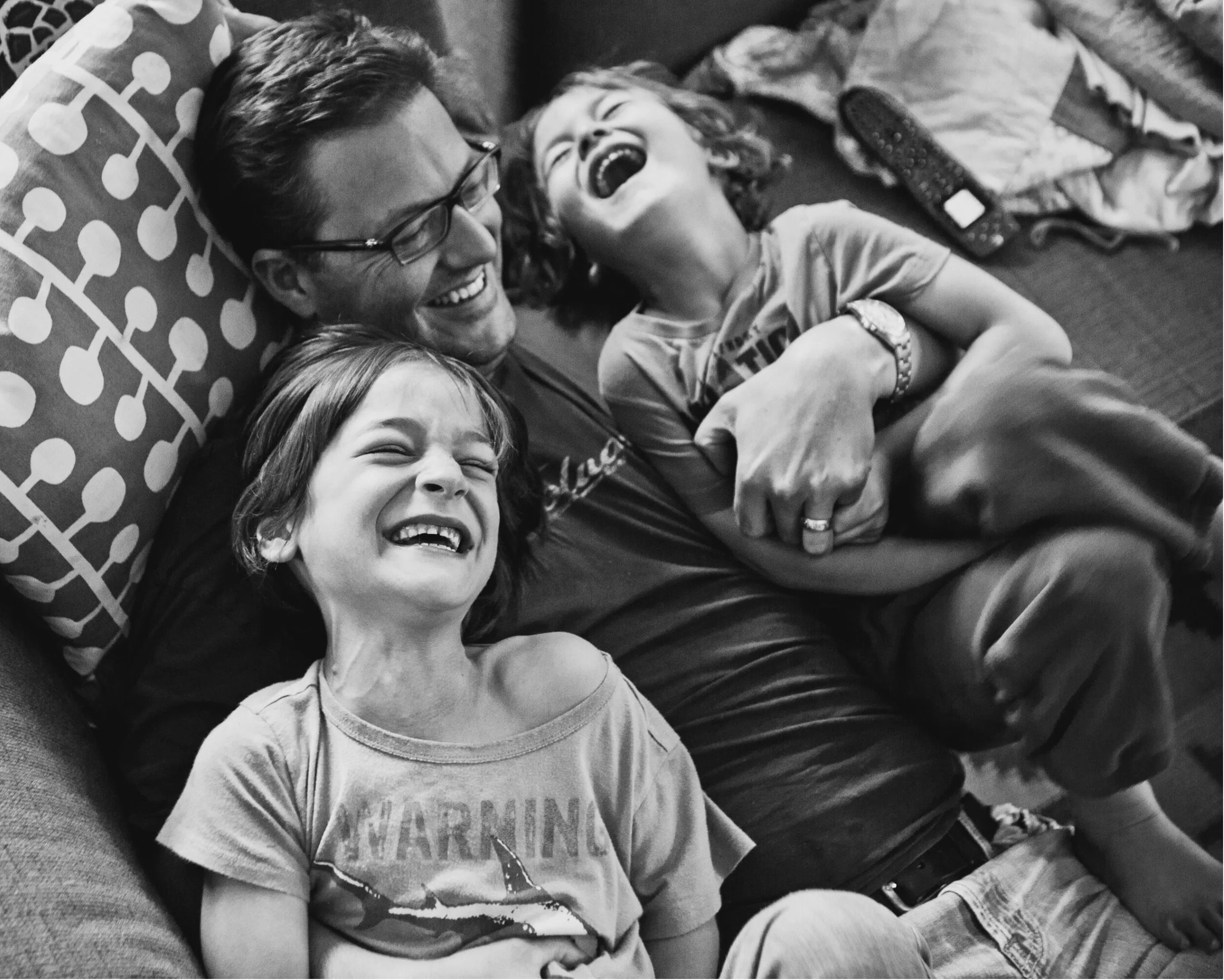A man and two children are lying on a couch, laughing and enjoying each other's company. The man is wearing glasses, and the children are wearing casual T-shirts, all captured in a joyful moment.