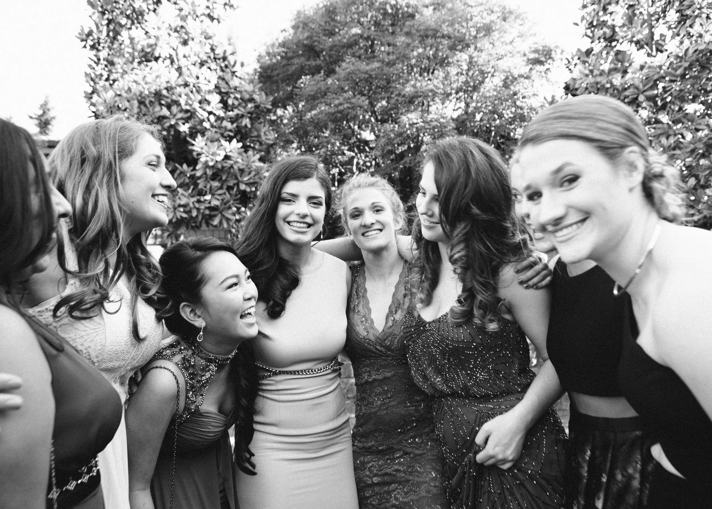 A group of smiling young women standing close together outdoors, with trees and sky in the background, celebrating and enjoying each other's company.
