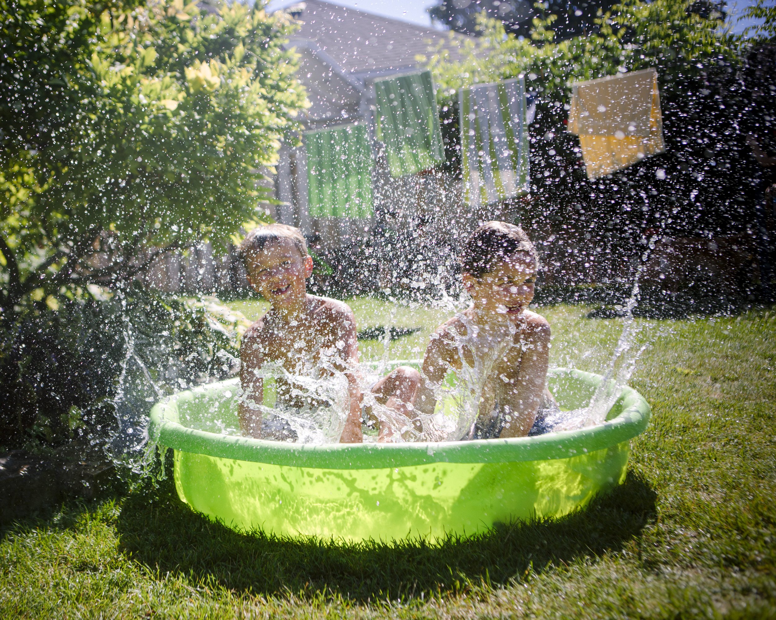 Two children playing in a green plastic pool while water splashes around them, outdoors in a backyard with green bushes and laundry hanging on a clothesline.