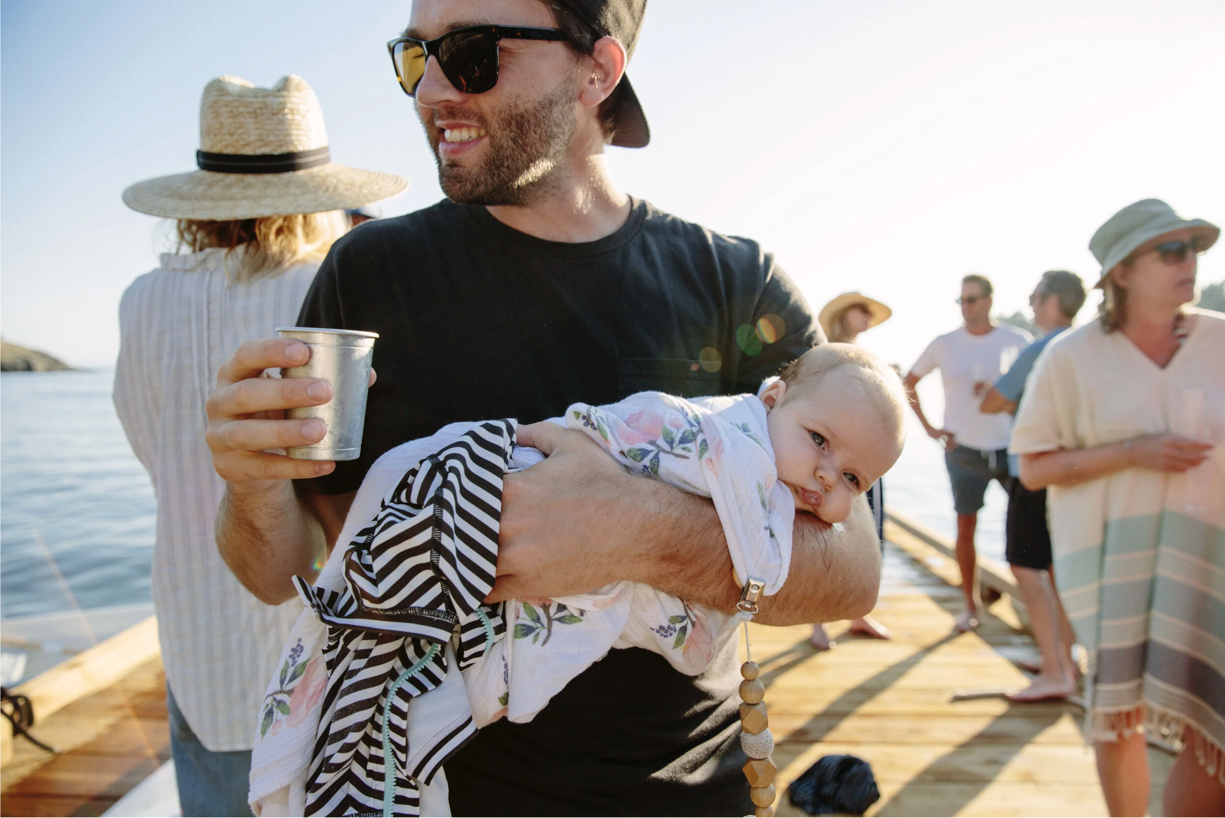 Man smiling and holding a baby at a seaside dock during sunset, with people in the background wearing summer clothes and sun hats.