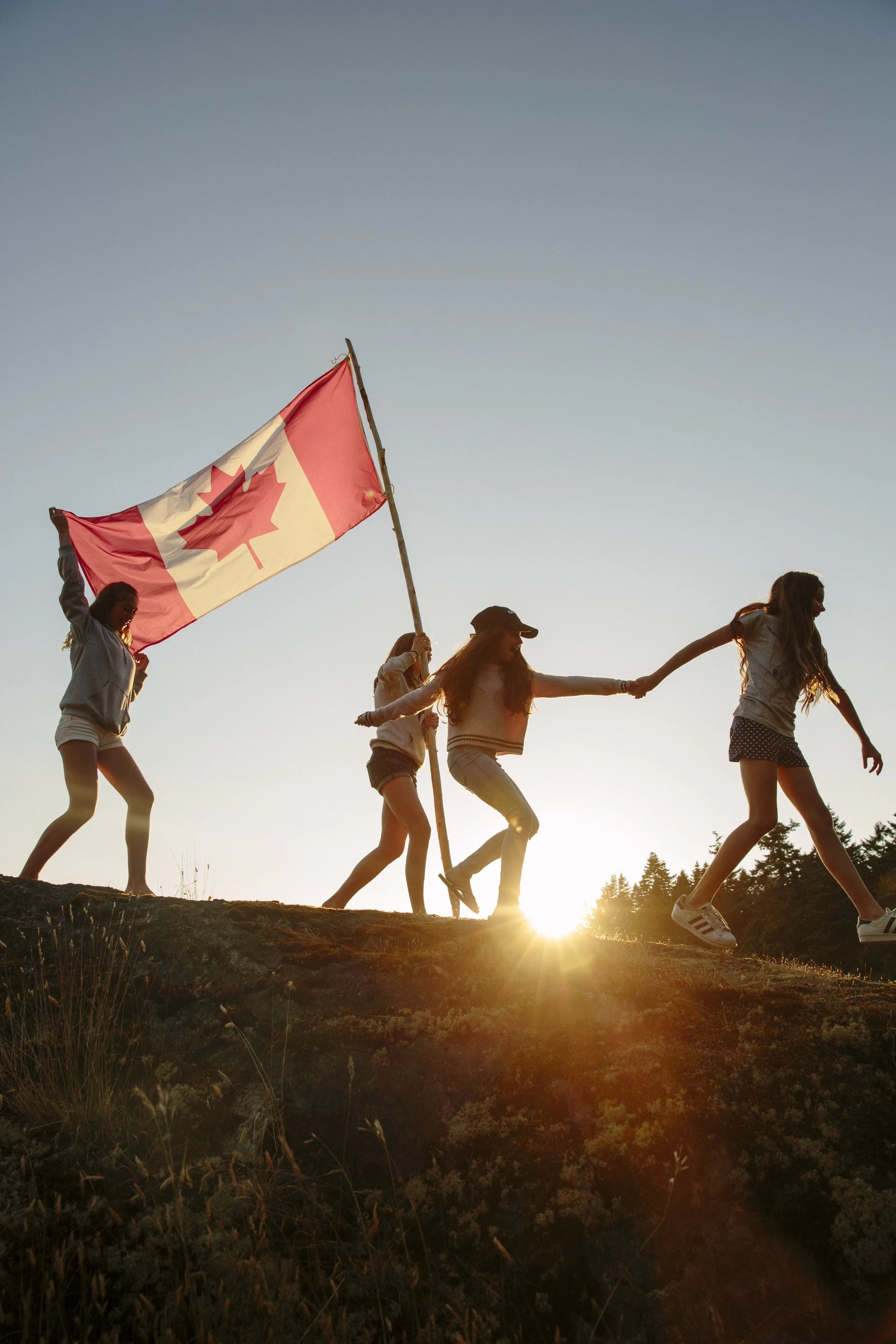 Group of young women holding hands and walking with a Canadian flag during sunset or sunrise in an outdoor natural setting.