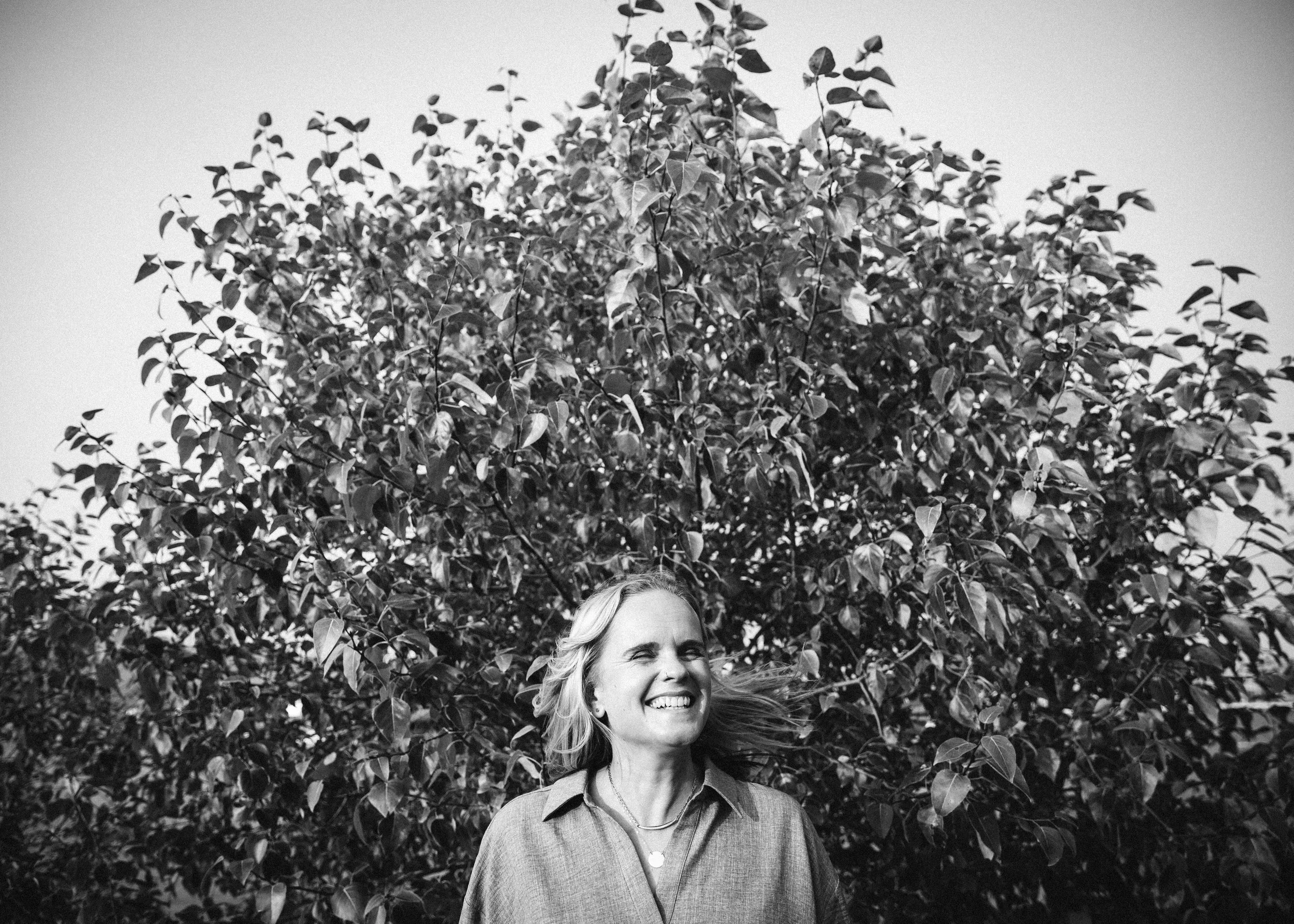 A woman with blonde hair smiling and looking to the side, standing in front of dense leafy tree branches, with part of her face and upper body visible in front of a clear sky.