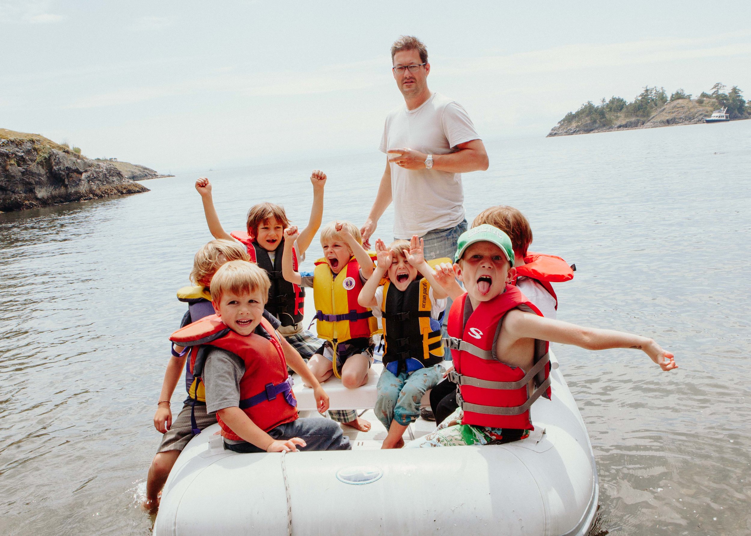 Children and an adult on a boat with life jackets, celebrating and enjoying on the water near a rocky shoreline and small island with trees.