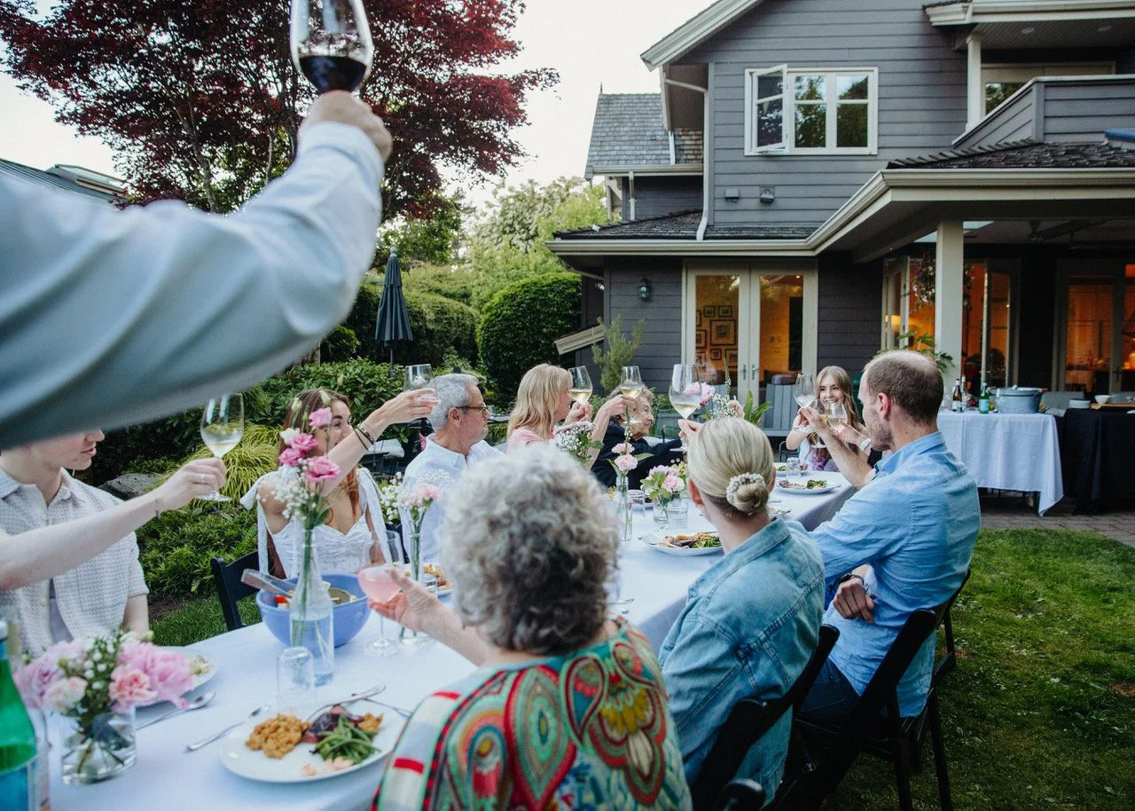 People gathered around a dining table outdoors at sunset, raising glasses in a toast during a celebration at a house with a garden and a patio.