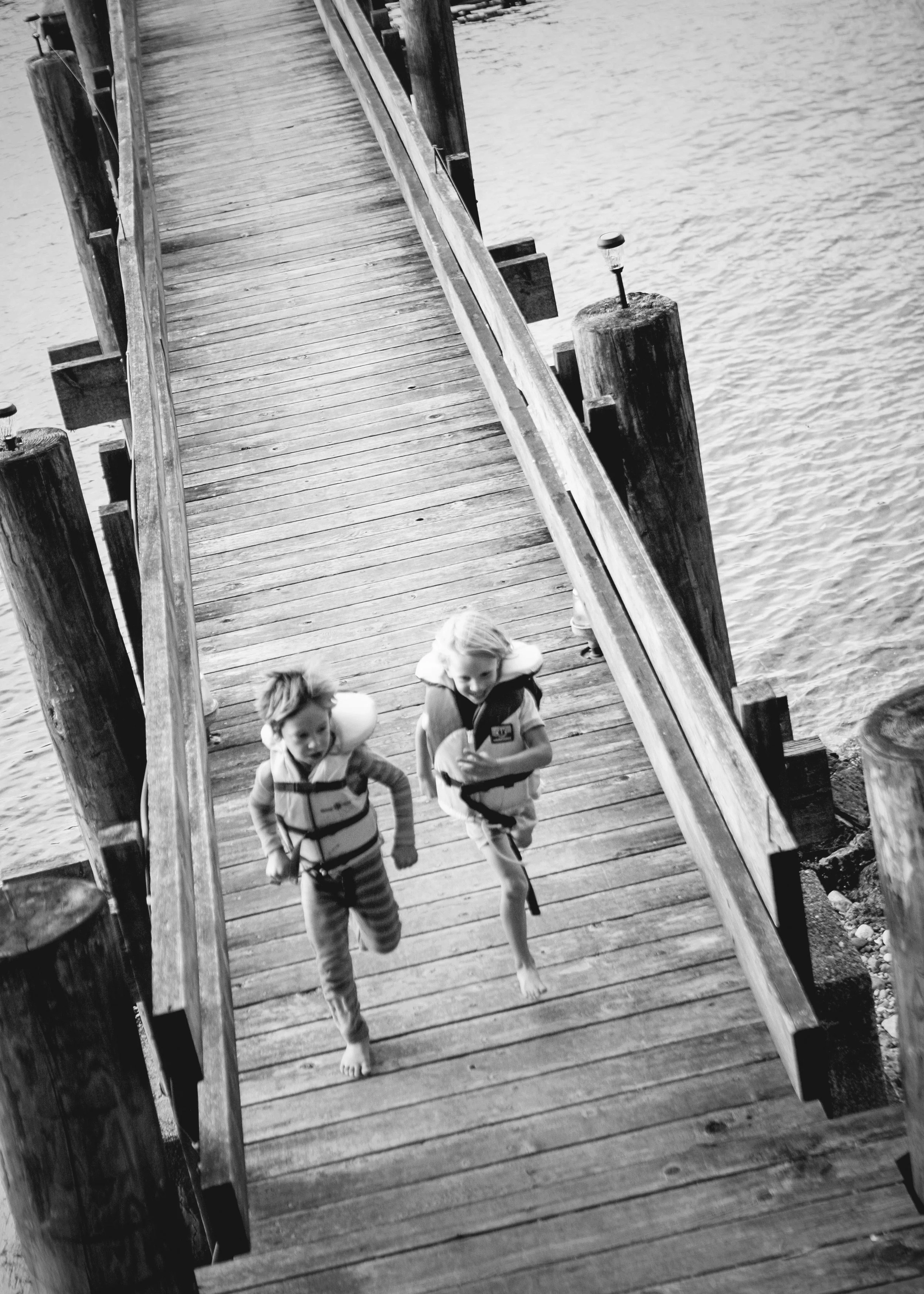 Two children walking on a wooden pier by the water, wearing life jackets.