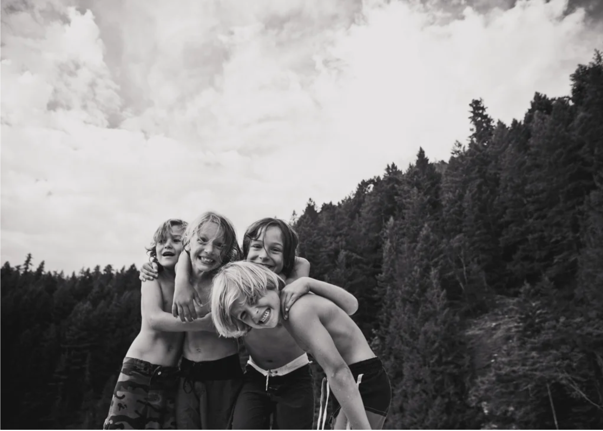 A group of smiling children with wet hair, embracing each other outdoors with a forested mountain and cloudy sky in the background.