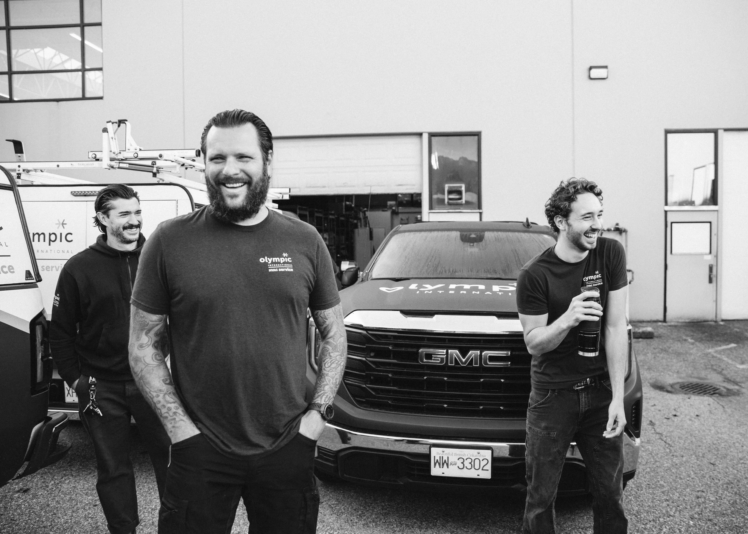Three smiling men standing in front of a GMC vehicle near a building with large windows, some of them wearing black t-shirts with a logo.