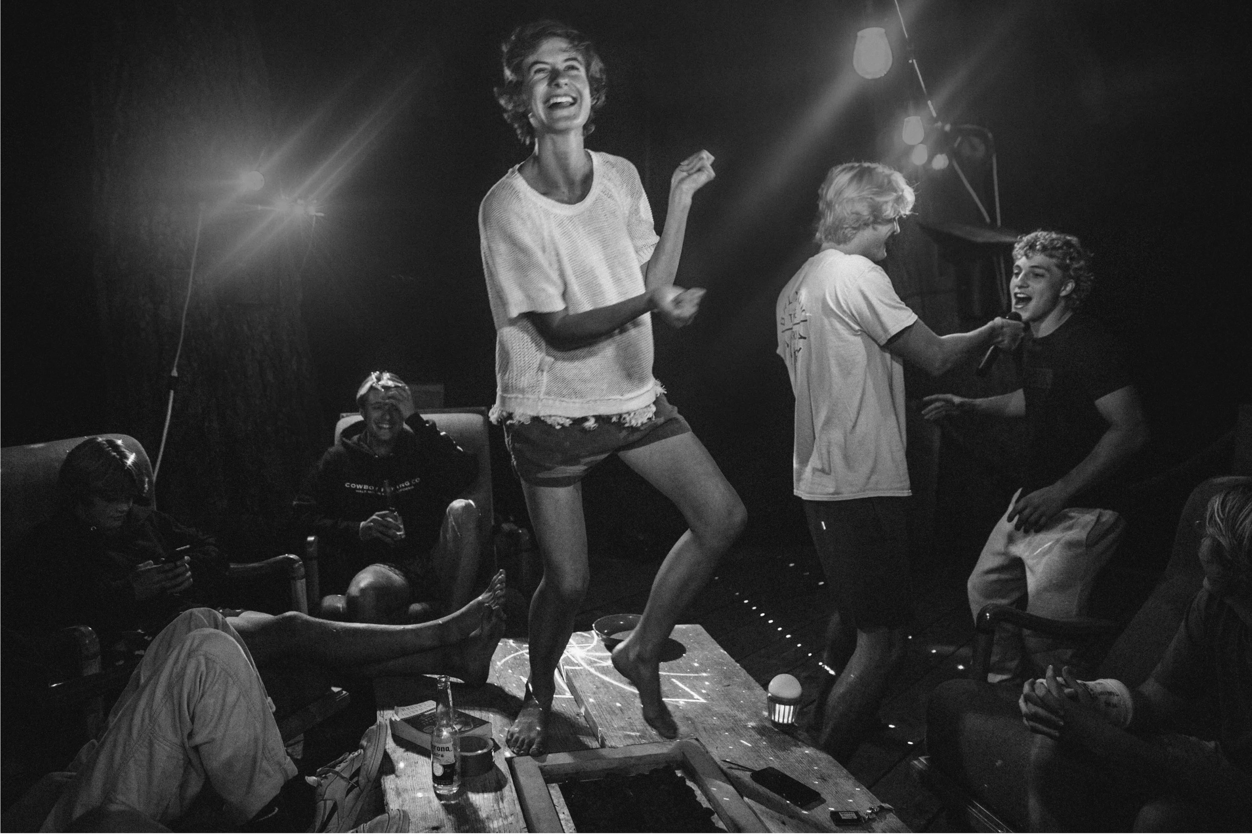 Black and white photo of a woman dancing on a wooden stage, smiling, with a microphone in her hand, while others sit and watch, with stage lights overhead.