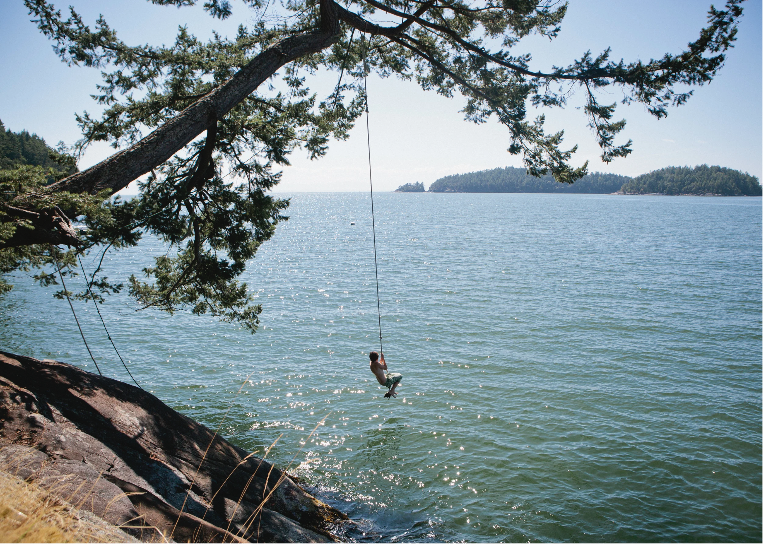 A boy on a swing hanging from a tree over a lake with distant islands under a clear blue sky.