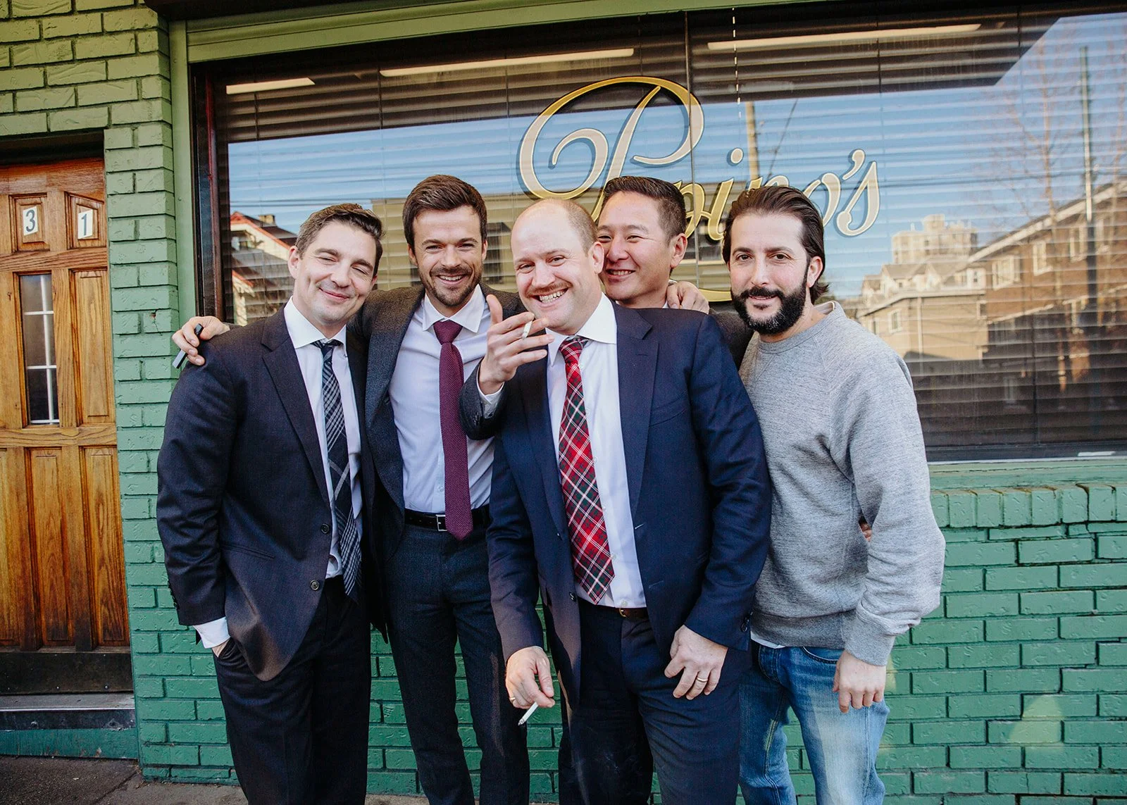 Group of five men standing outside a building with green brick wall, some in suits, smiling and posing for a photo.