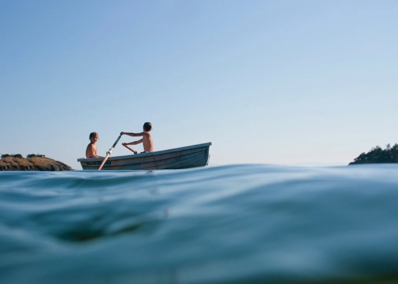 Two children in a small boat on calm water with a clear blue sky and a distant shoreline.