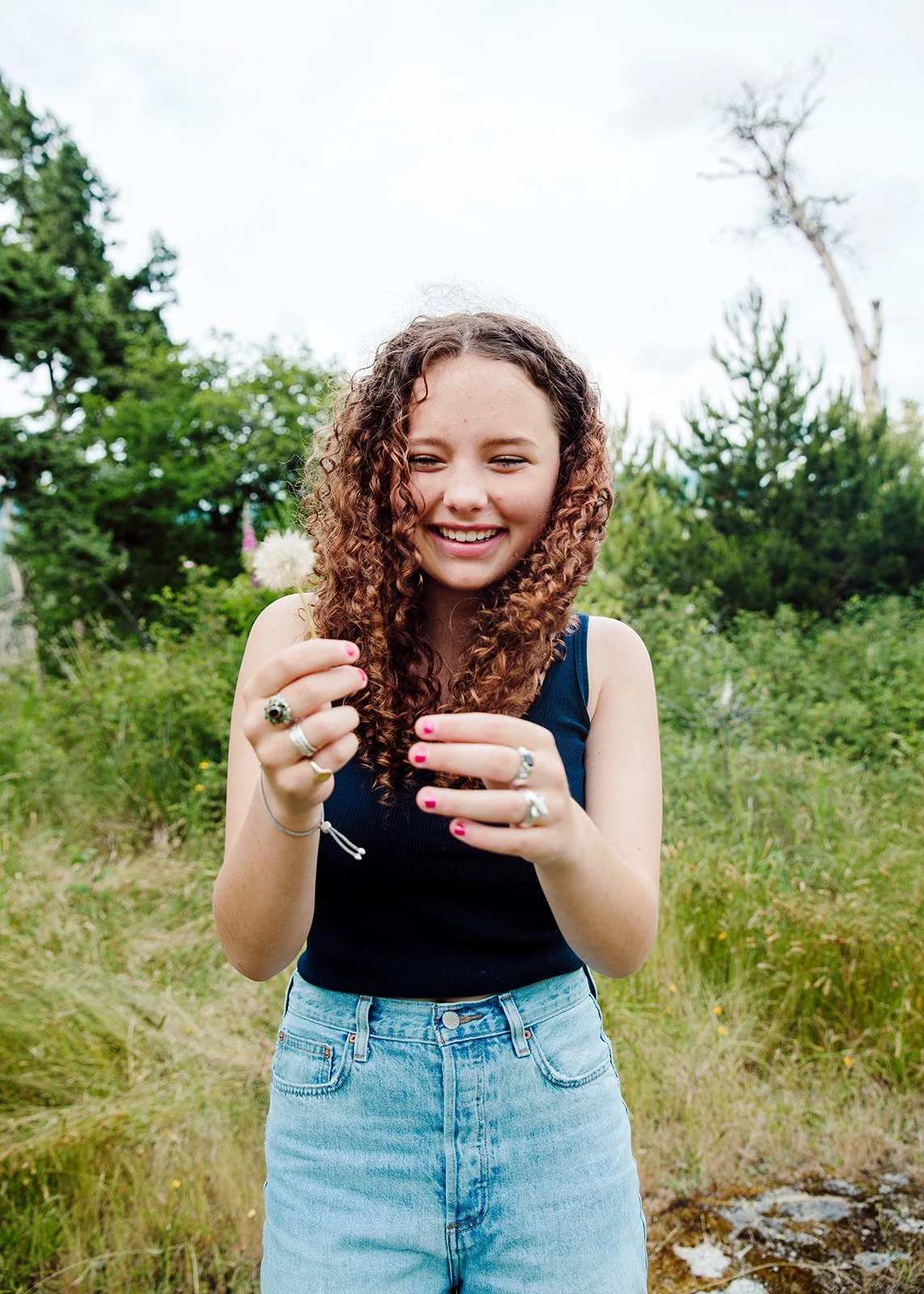 A young woman with curly brown hair smiling and holding a dandelion flower outdoors in a green, natural setting.