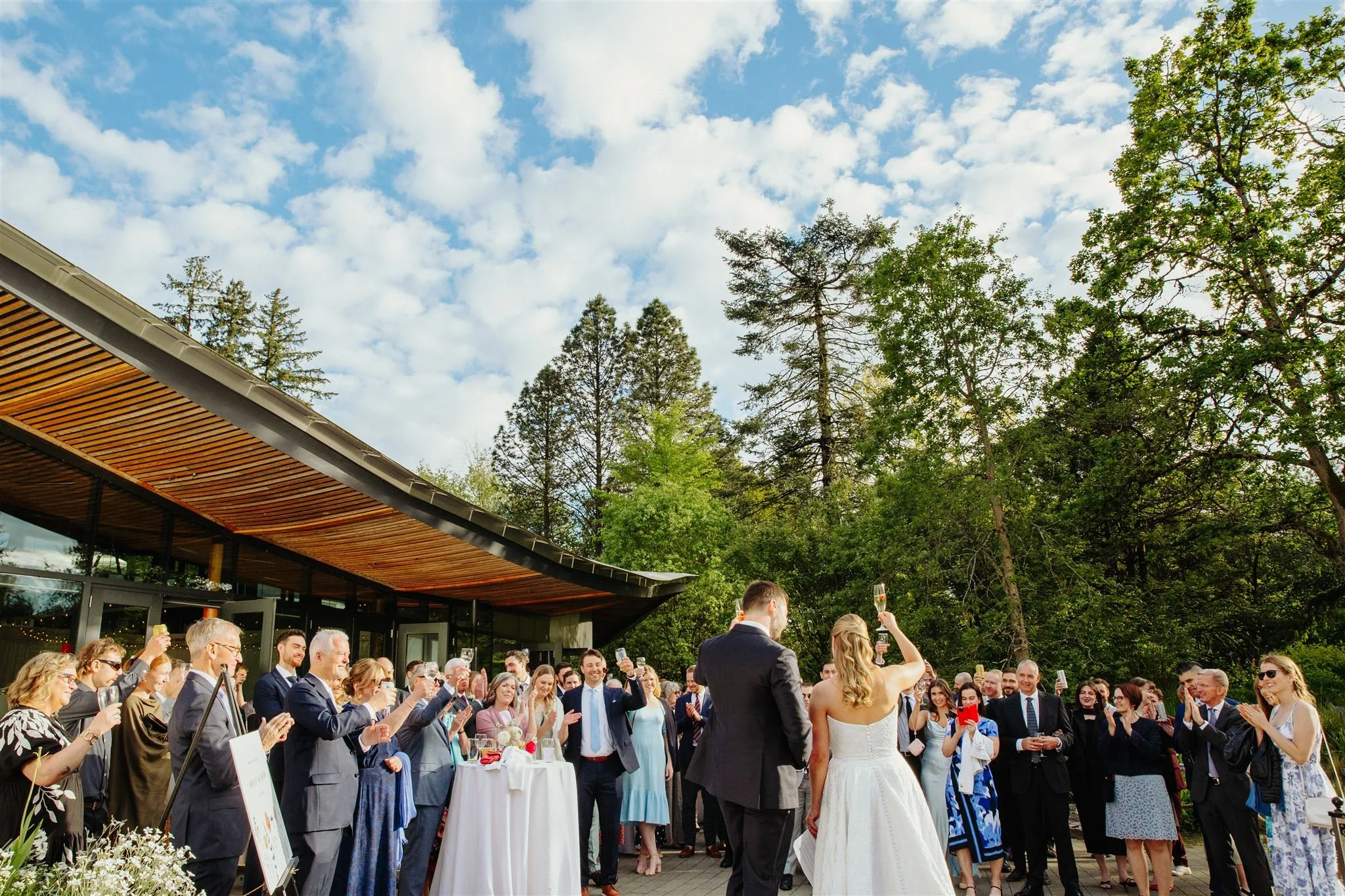 A wedding celebration outdoors with a bride and groom raising glasses, surrounded by guests clapping and smiling against a backdrop of green trees and a partly cloudy sky.