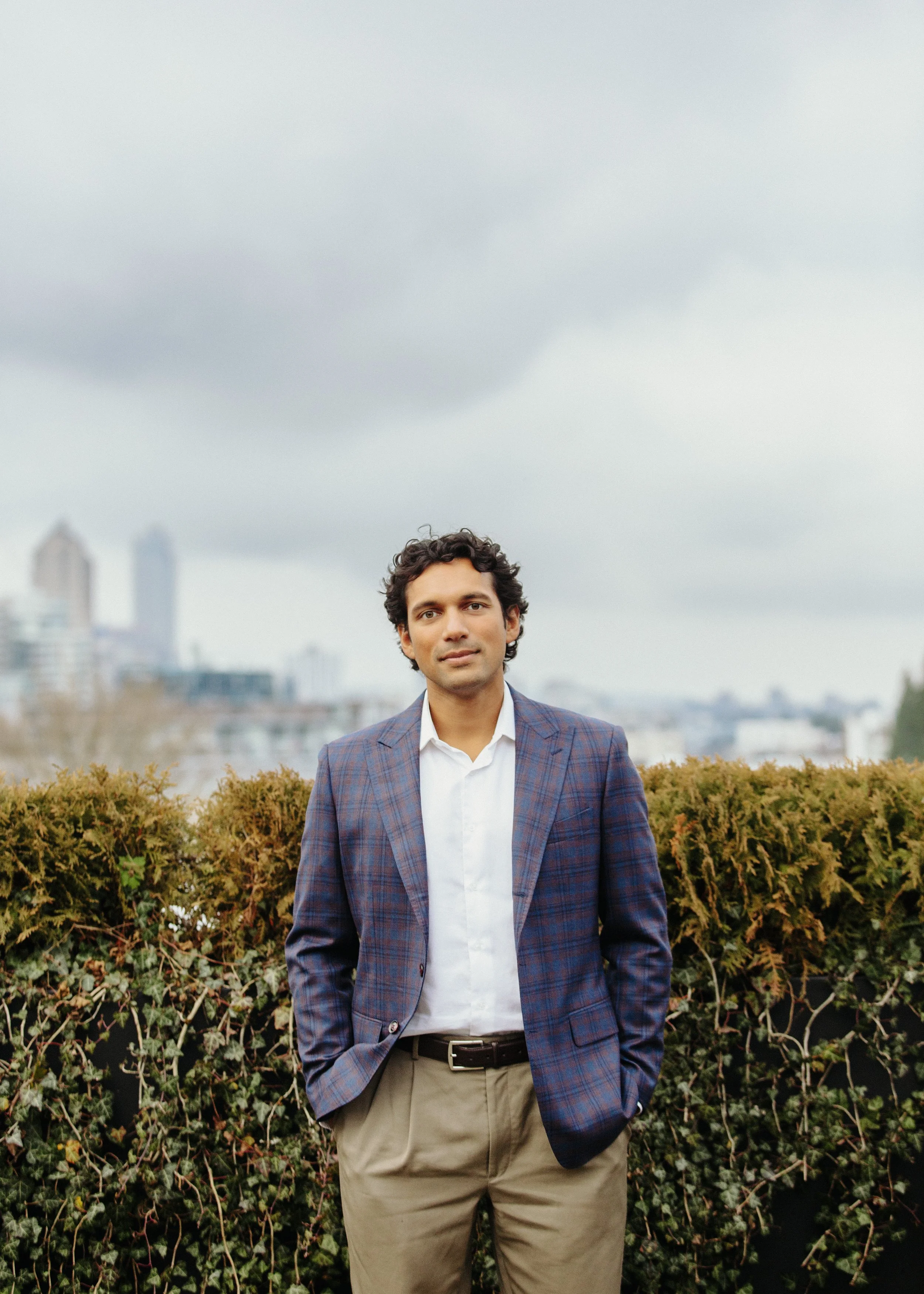 A man wearing a checked blazer and beige pants standing outdoors in front of a hedge on a city rooftop with skyscrapers in the background under a cloudy sky.
