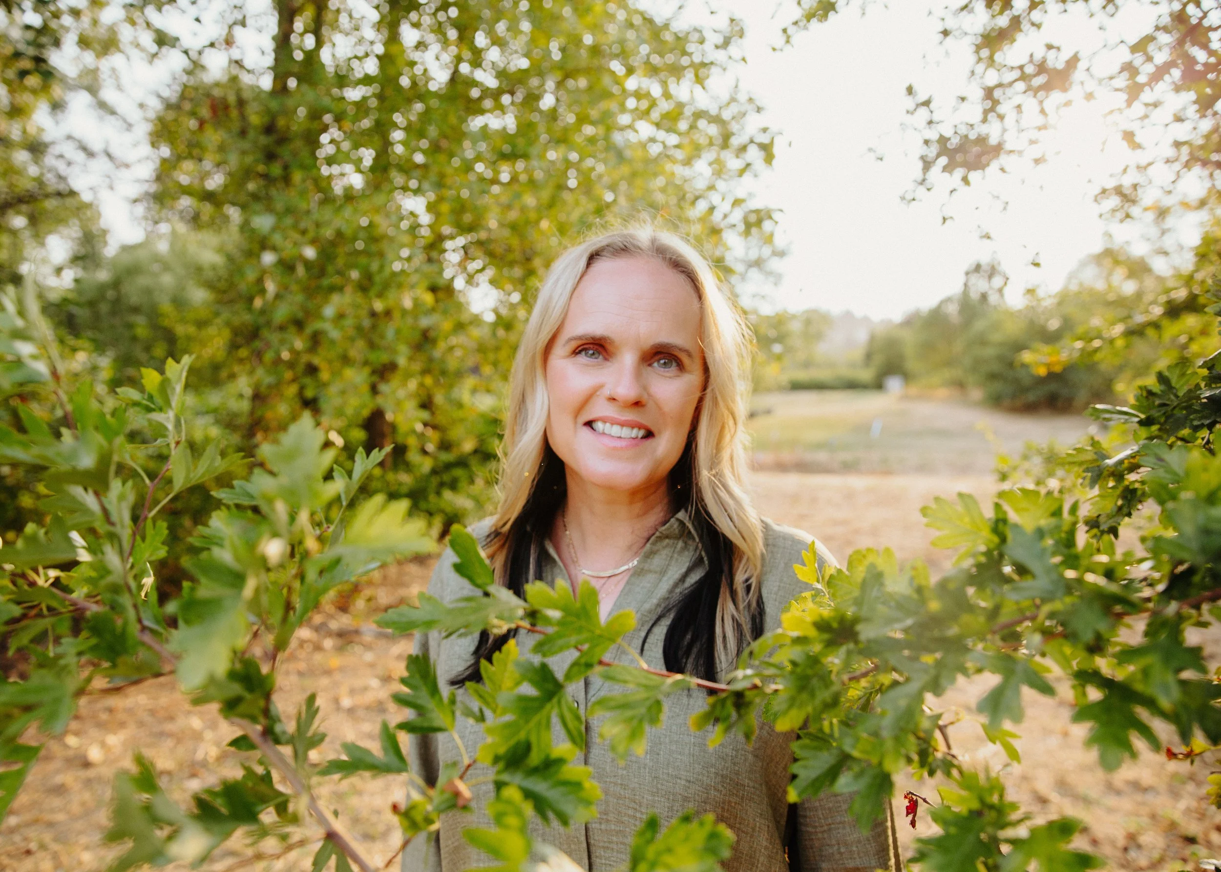 A woman with blonde hair smiling in an outdoor setting surrounded by green bushes and trees during daytime.