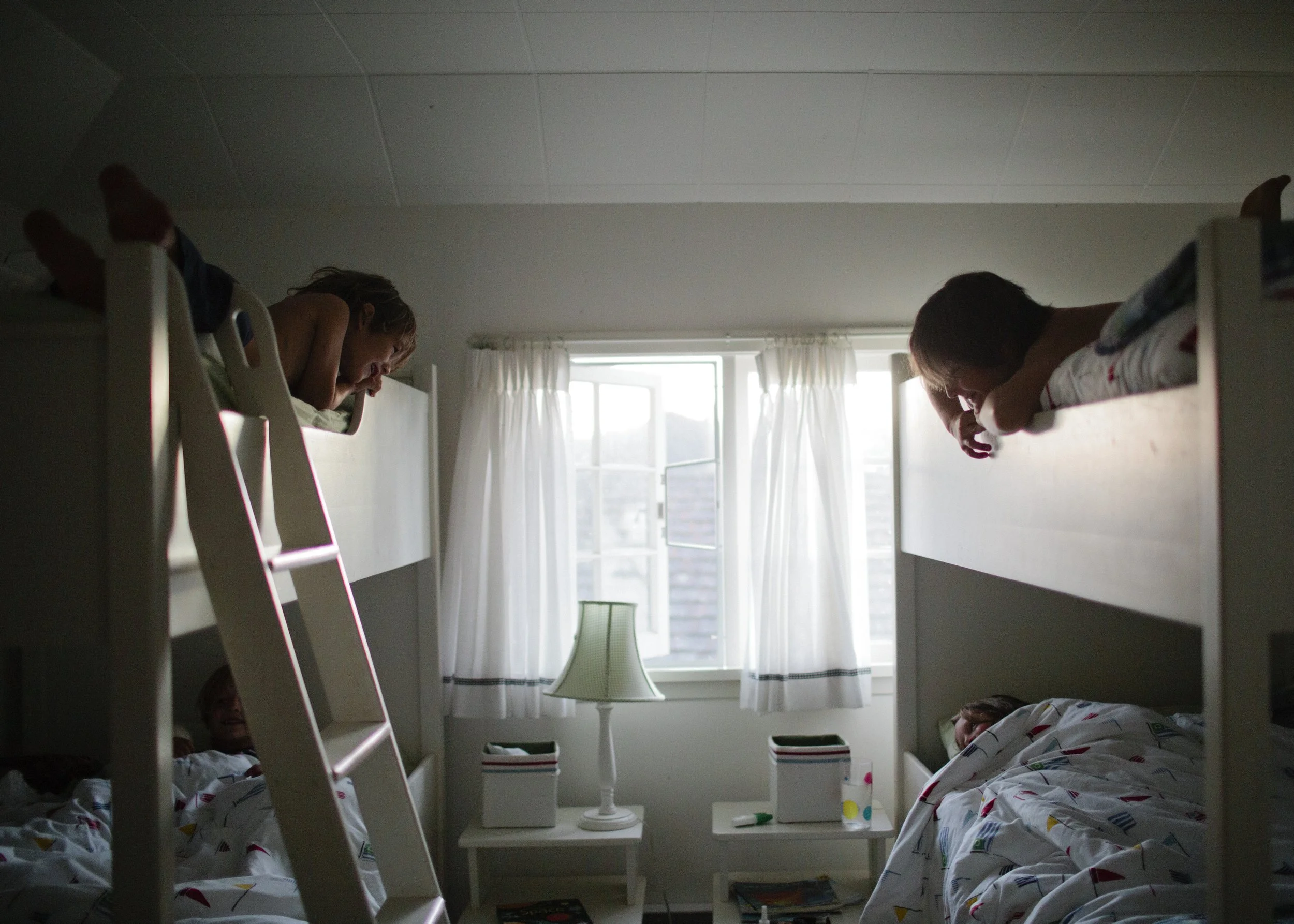 Children sleeping in a dormitory room with bunk beds, a window, white curtains, and a bedside table.