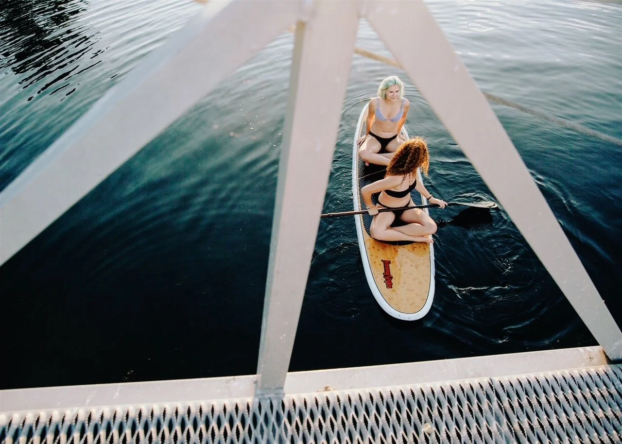 Two women on a paddleboard in the water, viewed from a bridge above, with a metal railing in the foreground.