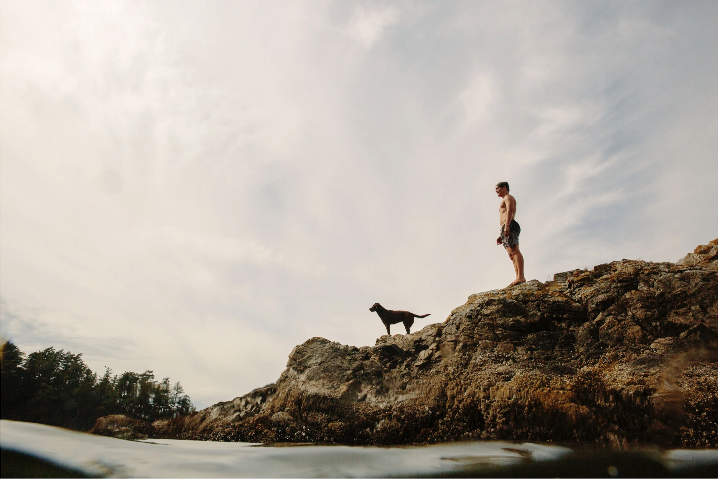 A shirtless man standing on a rocky ledge by the ocean with two dogs, one in front and one behind, under a cloudy sky.