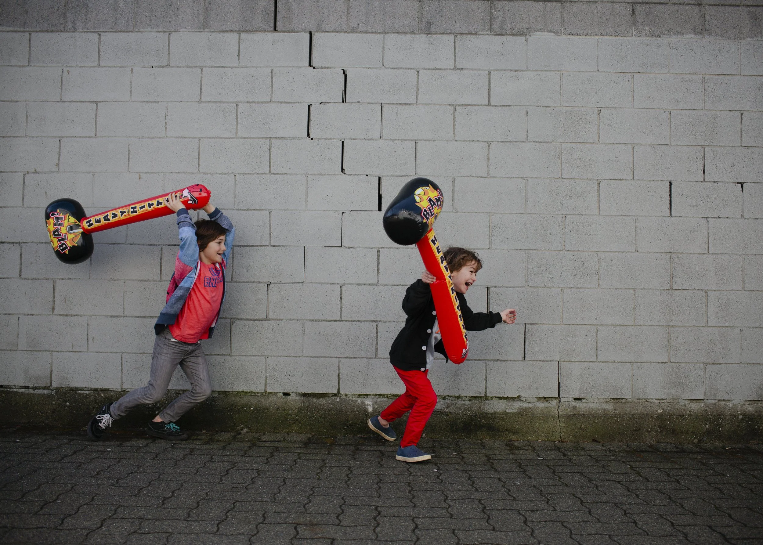 Two children playing outside with inflatable hammer toys, one child chasing behind and laughing, against a concrete block wall.