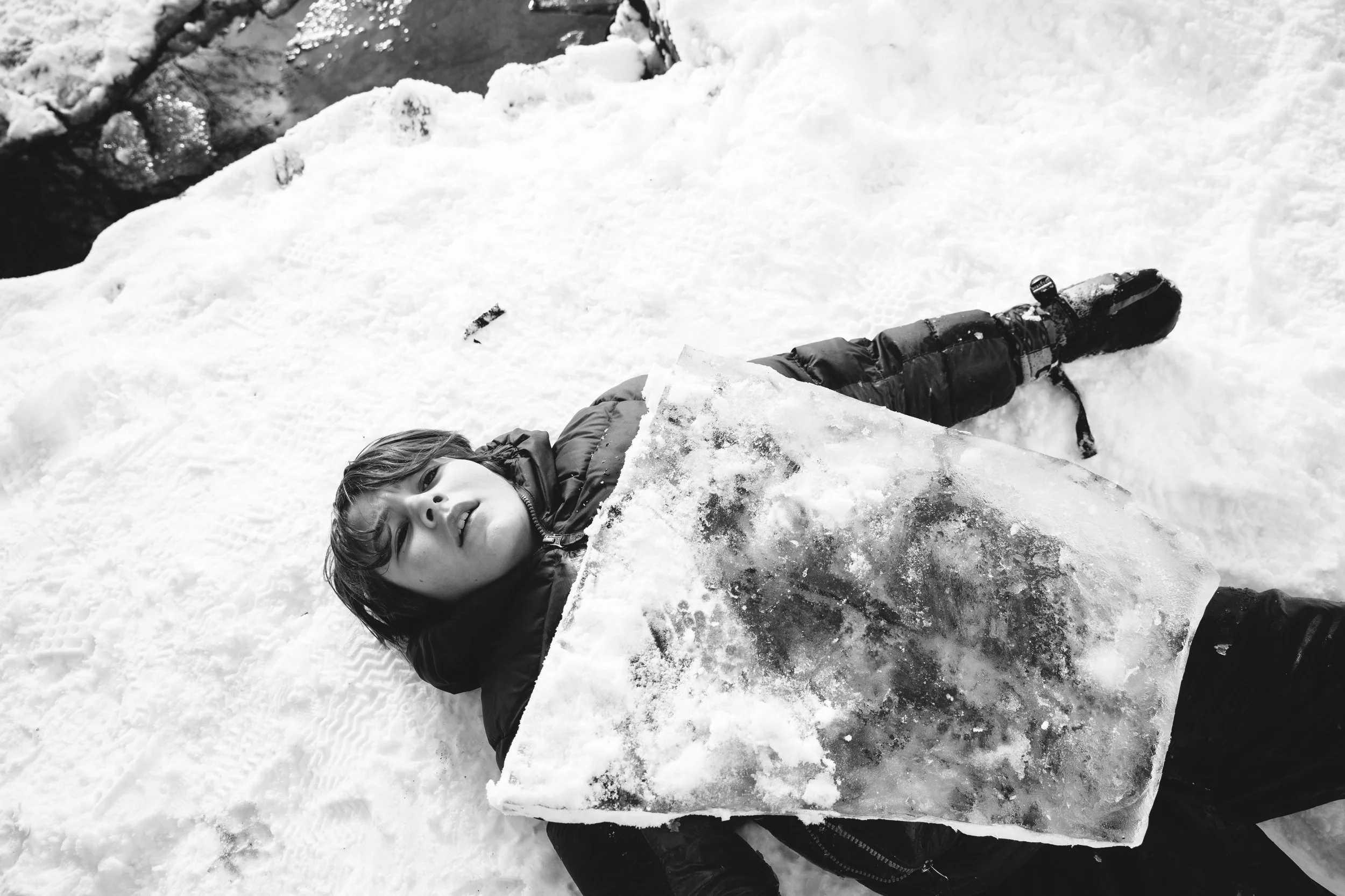A young person lying on the snow holding a large piece of ice, with snow-covered ground and some dark water patches in the background.