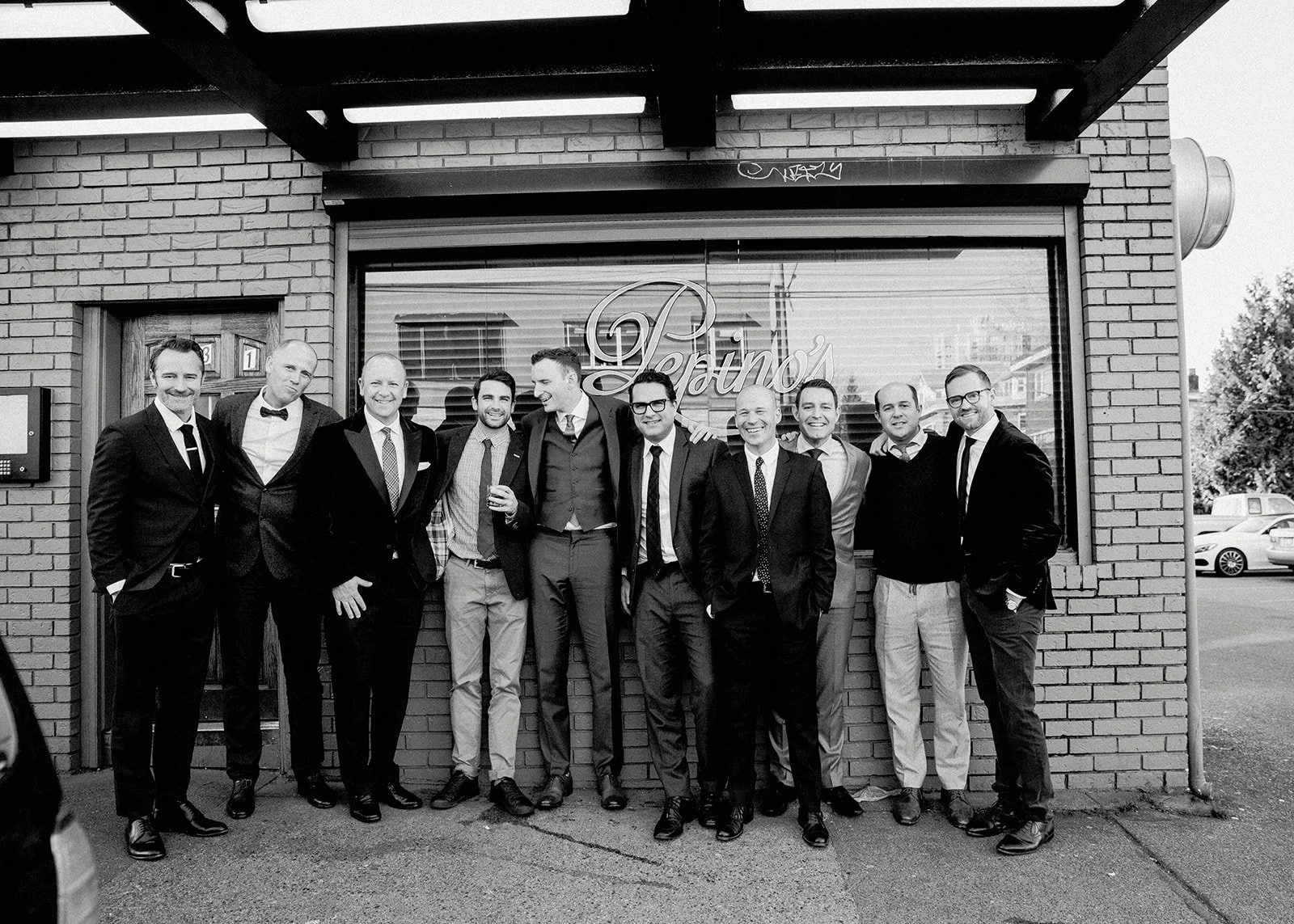 Group of eleven men in suits standing outside a brick building with large window, smiling for a photo.