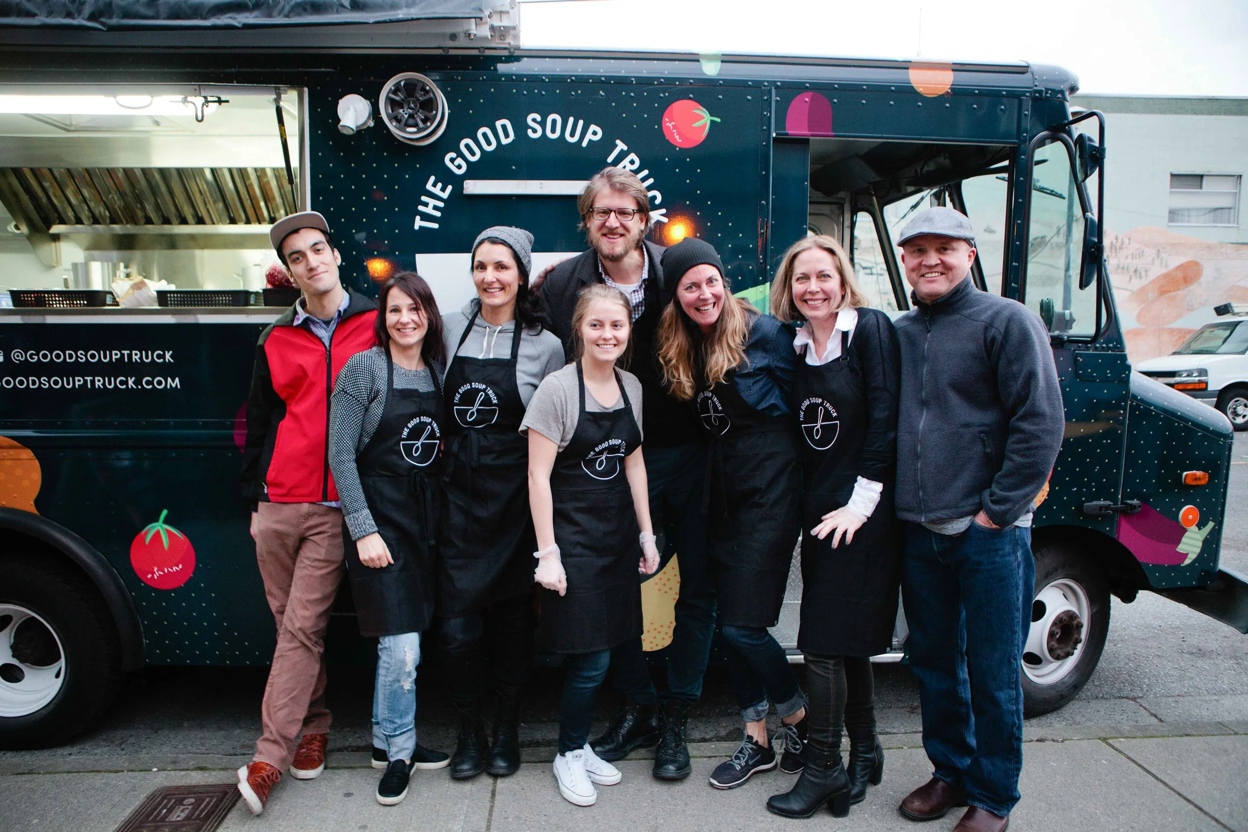 Group of eight people standing in front of a food truck labeled "The Good Soup Truck." The group includes five women and three men, smiling and posing for the photo, some wearing black aprons with the truck's logo. The truck has colorful graphics and
