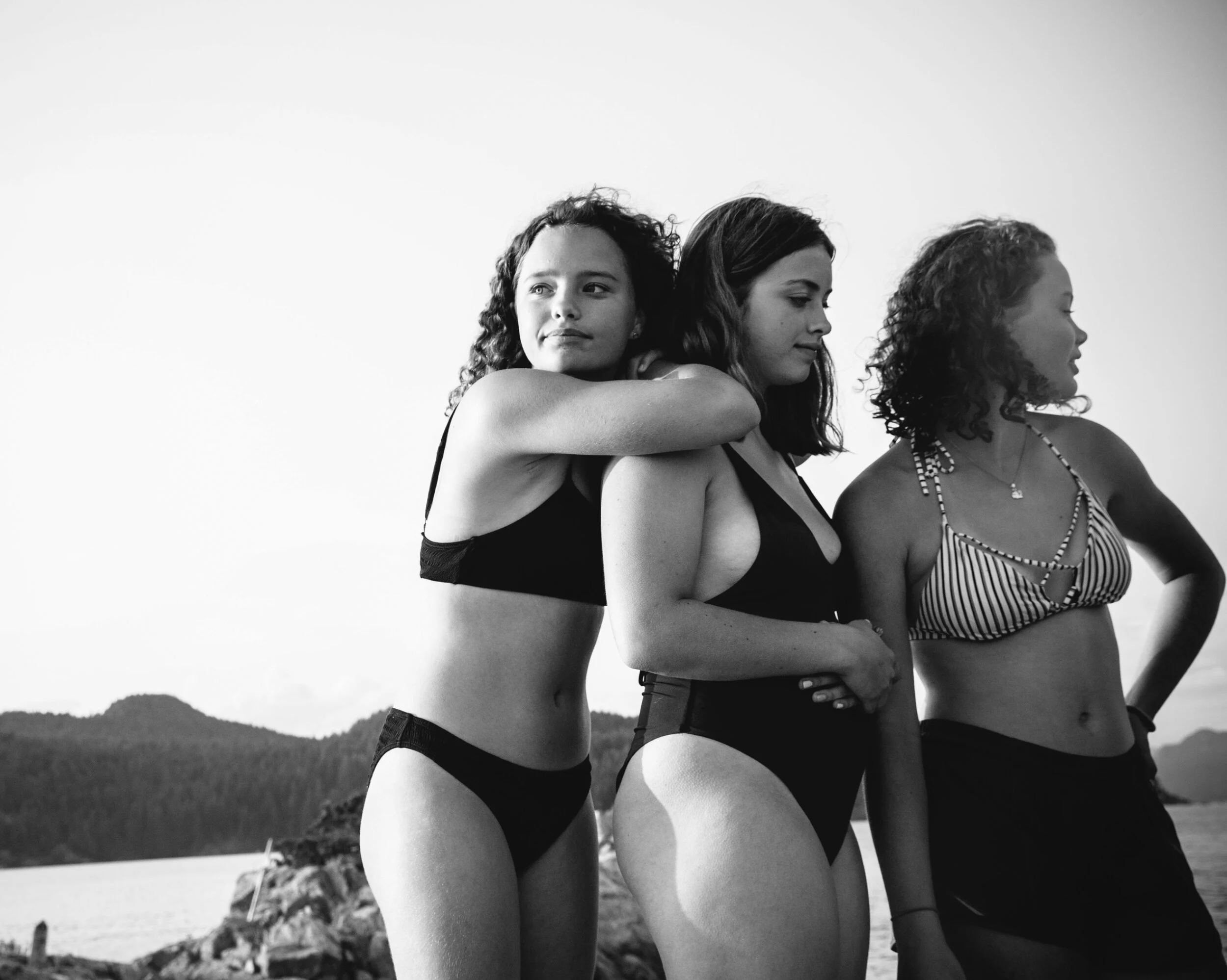 Three women standing on a beach with mountains and water in the background, wearing swimsuits, one hugging the others from behind.