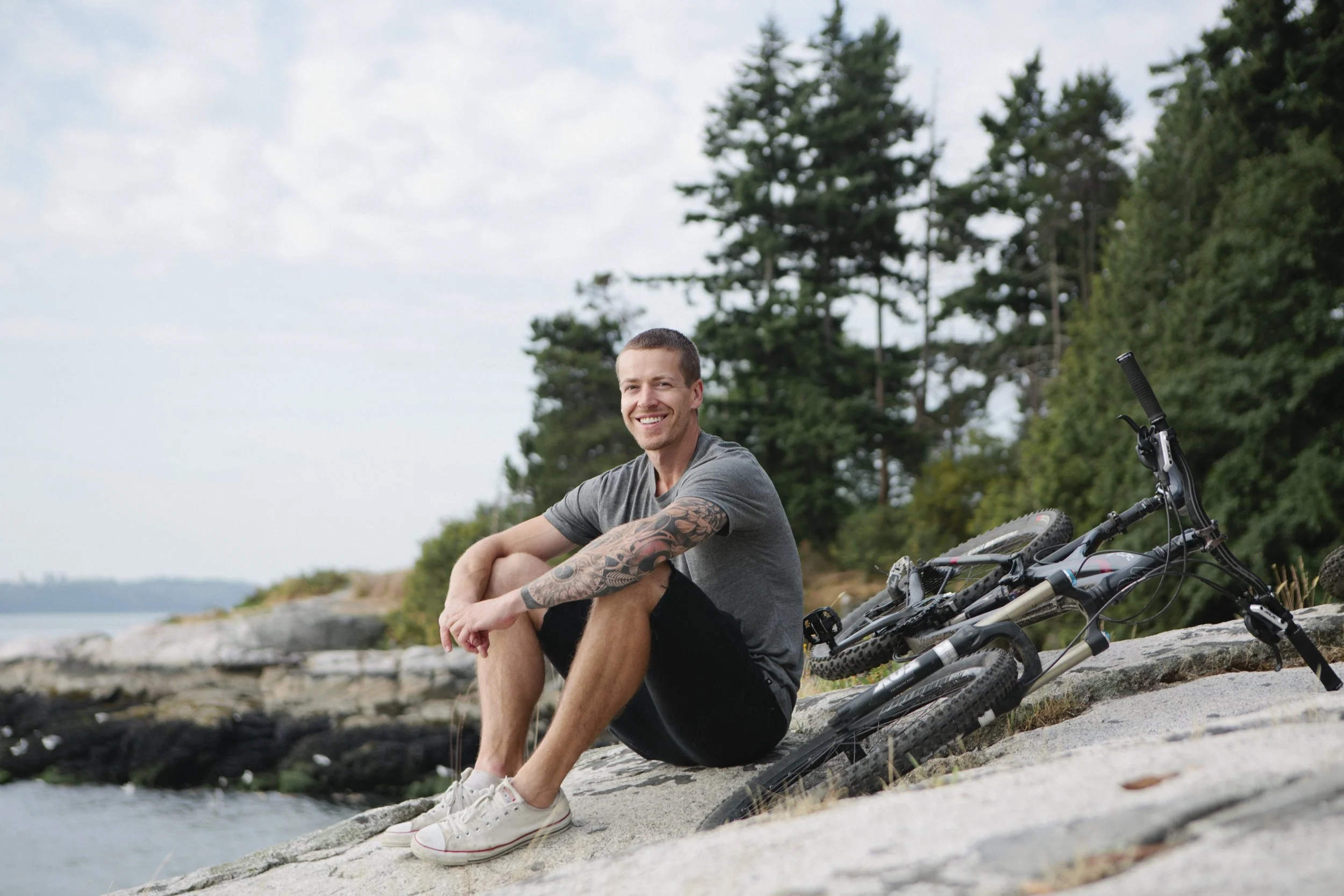A man sitting on a rocky shoreline by the water, smiling, with a mountain bike lying on the ground next to him, surrounded by trees under a partly cloudy sky.