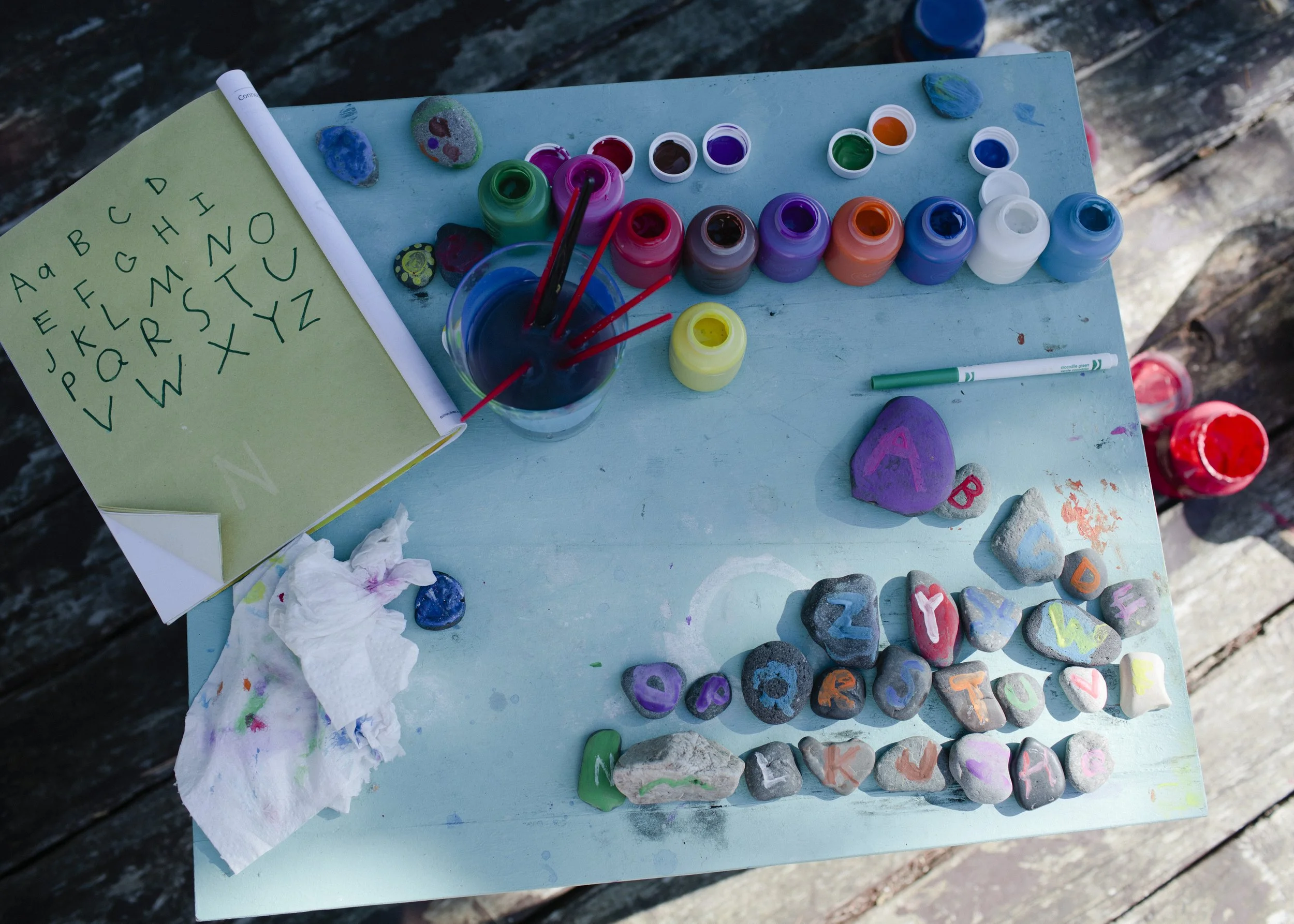 Paint bottles, rocks, and painted stones on a blue table, next to a green notebook with alphabet letters.