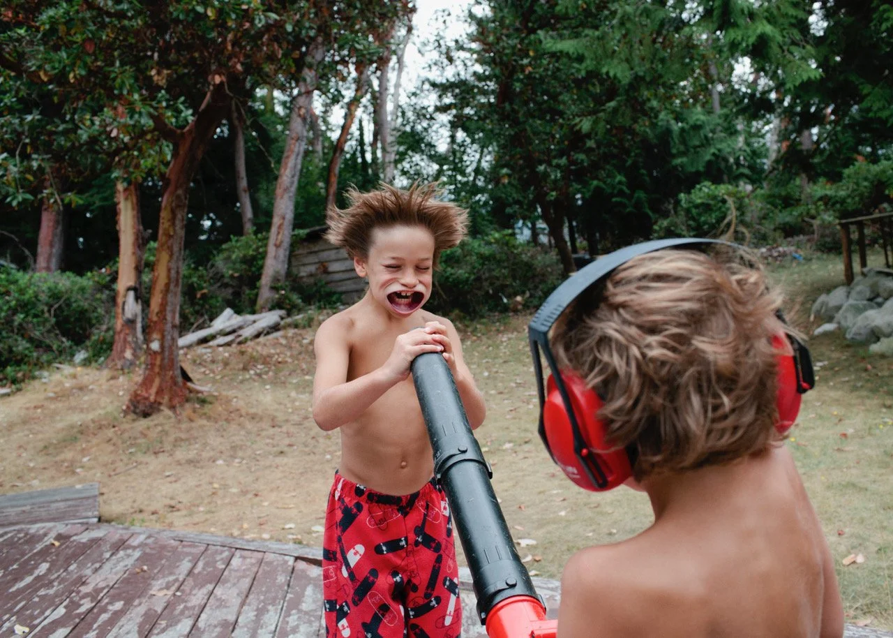 Two young boys are outside in a wooded yard, one with a hairpiece and no shirt, laughing and holding a leaf blower, while the other boy also shirtless, wears red ear protection and faces away.
